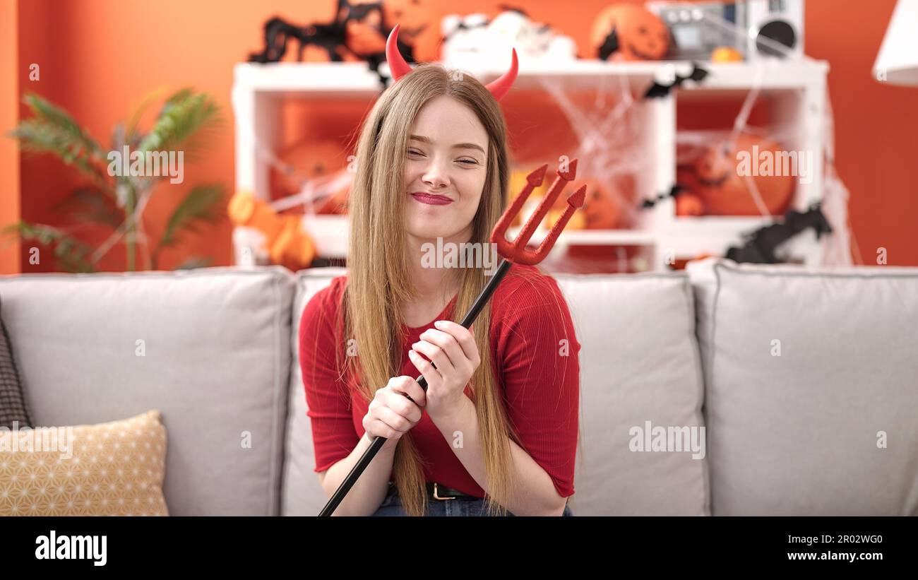 Giovane donna bionda sorridente indossando il costume diavolo seduto sul divano a casa Foto Stock