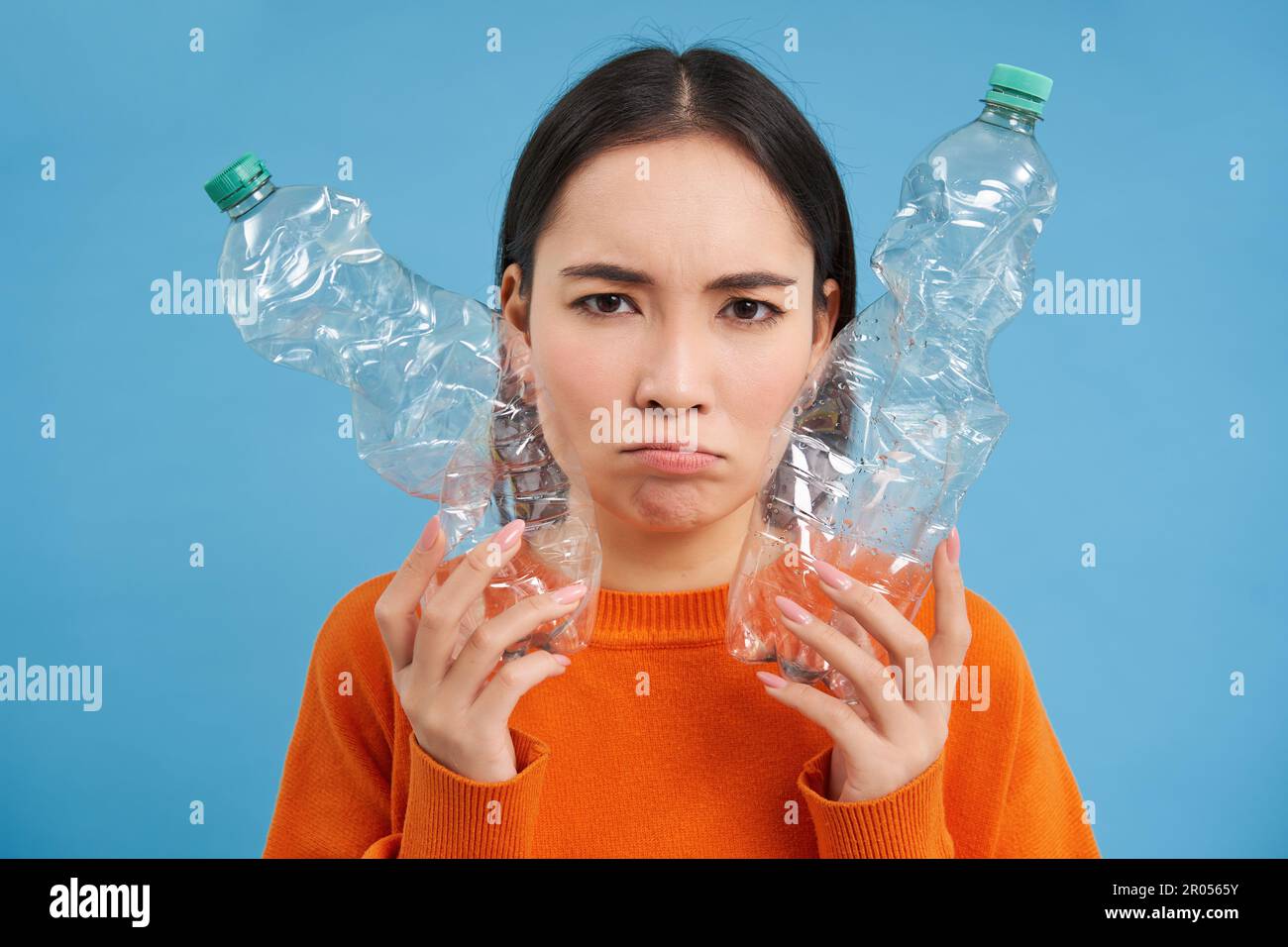 Ritratto di triste donna asiatica con bottiglie di plastica, sconvolto dalla mancanza di centri di riciclaggio, sfondo blu. Foto Stock
