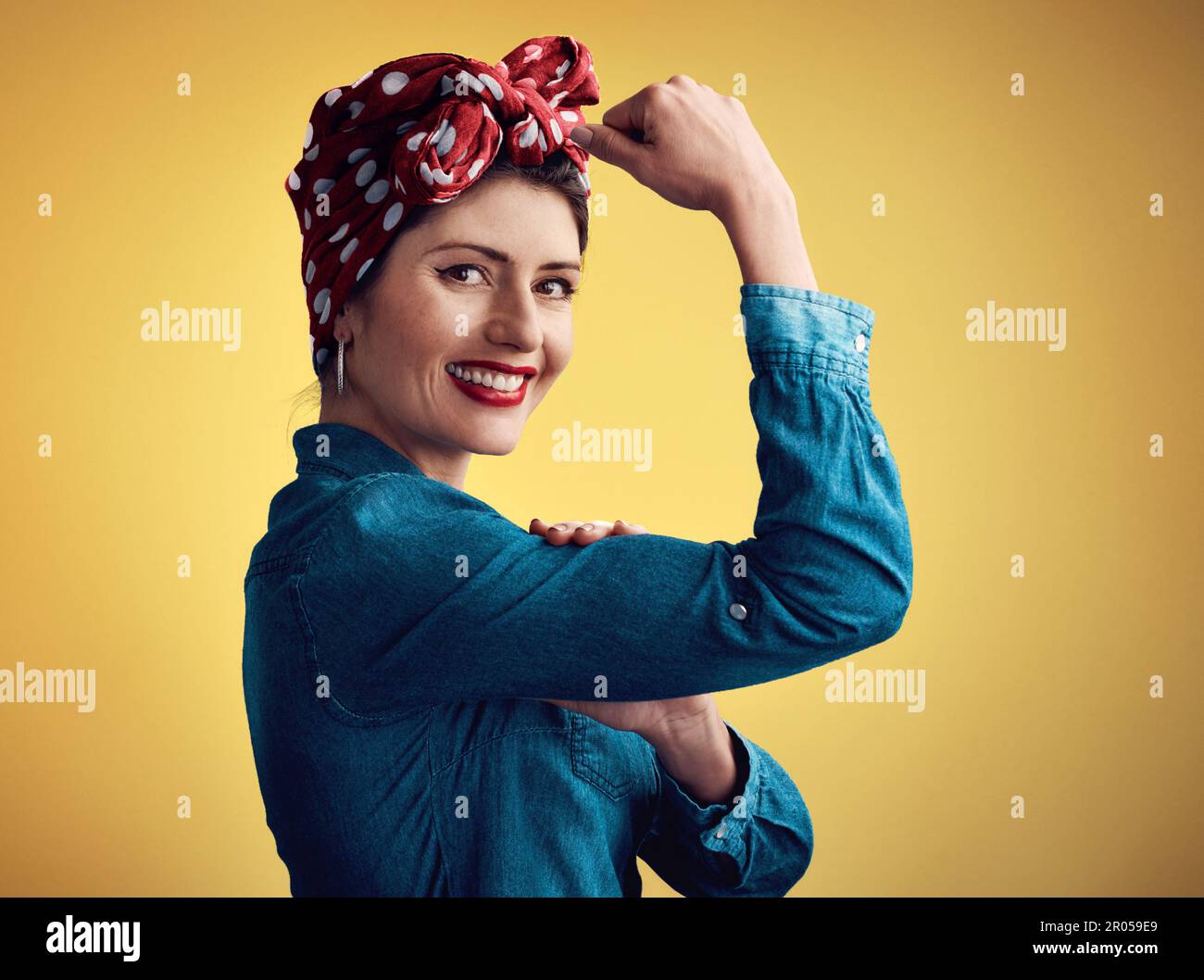 Femminismo in piena forza. Studio ritratto di una giovane donna attraente flexing il suo bicep mentre in piedi su uno sfondo giallo. Foto Stock