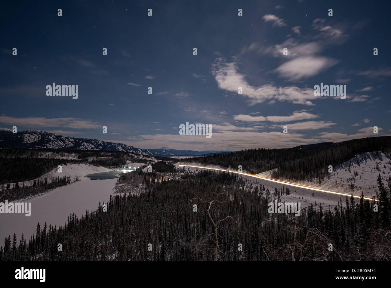 Scena notturna di una strada invernale nel Canada settentrionale, territorio dello Yukon. Preso sulla notte illuminata dalla luna con cielo stellato blu luminoso, abete rosso, alberi che circondano. Foto Stock