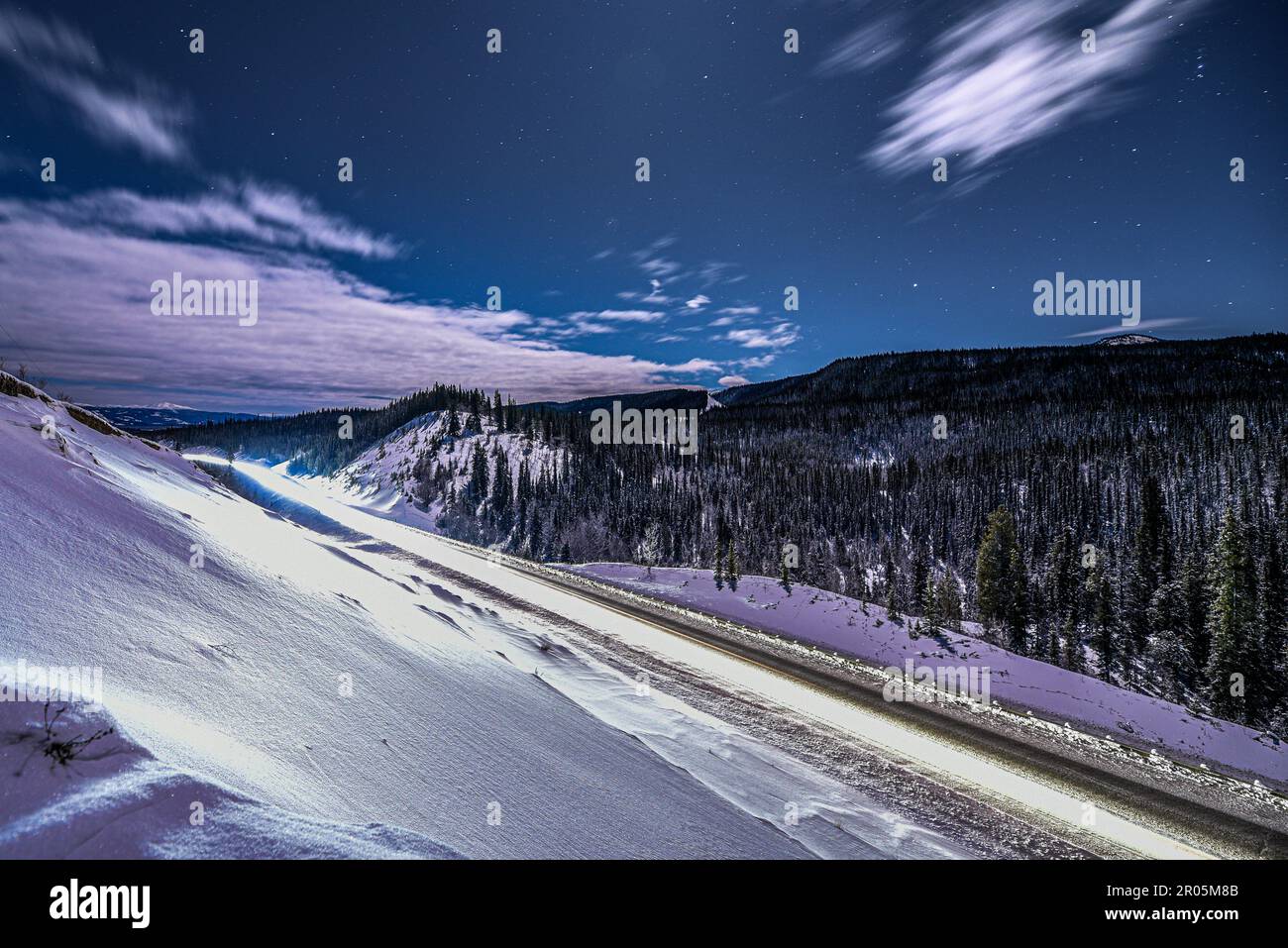 Scena notturna di una strada invernale nel Canada settentrionale, territorio dello Yukon. Preso sulla notte illuminata dalla luna con cielo stellato blu luminoso, abete rosso, alberi che circondano. Foto Stock