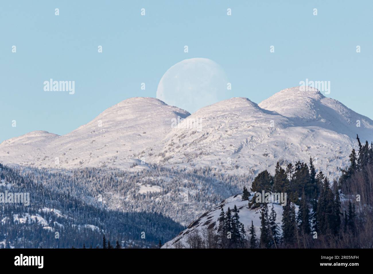 Luna che si affaccia su splendide montagne innevate nel nord del Canada durante l'inverno con cielo blu brillante e lago ghiacciato. Paesaggio, paesaggio. Foto Stock