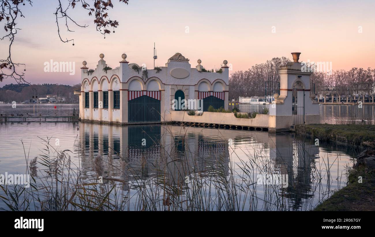 La tranquilla bellezza del lago Banyoles si riflette nel cielo al tramonto, incorniciato da una maestosa struttura costruita e circondato da alberi. Foto Stock