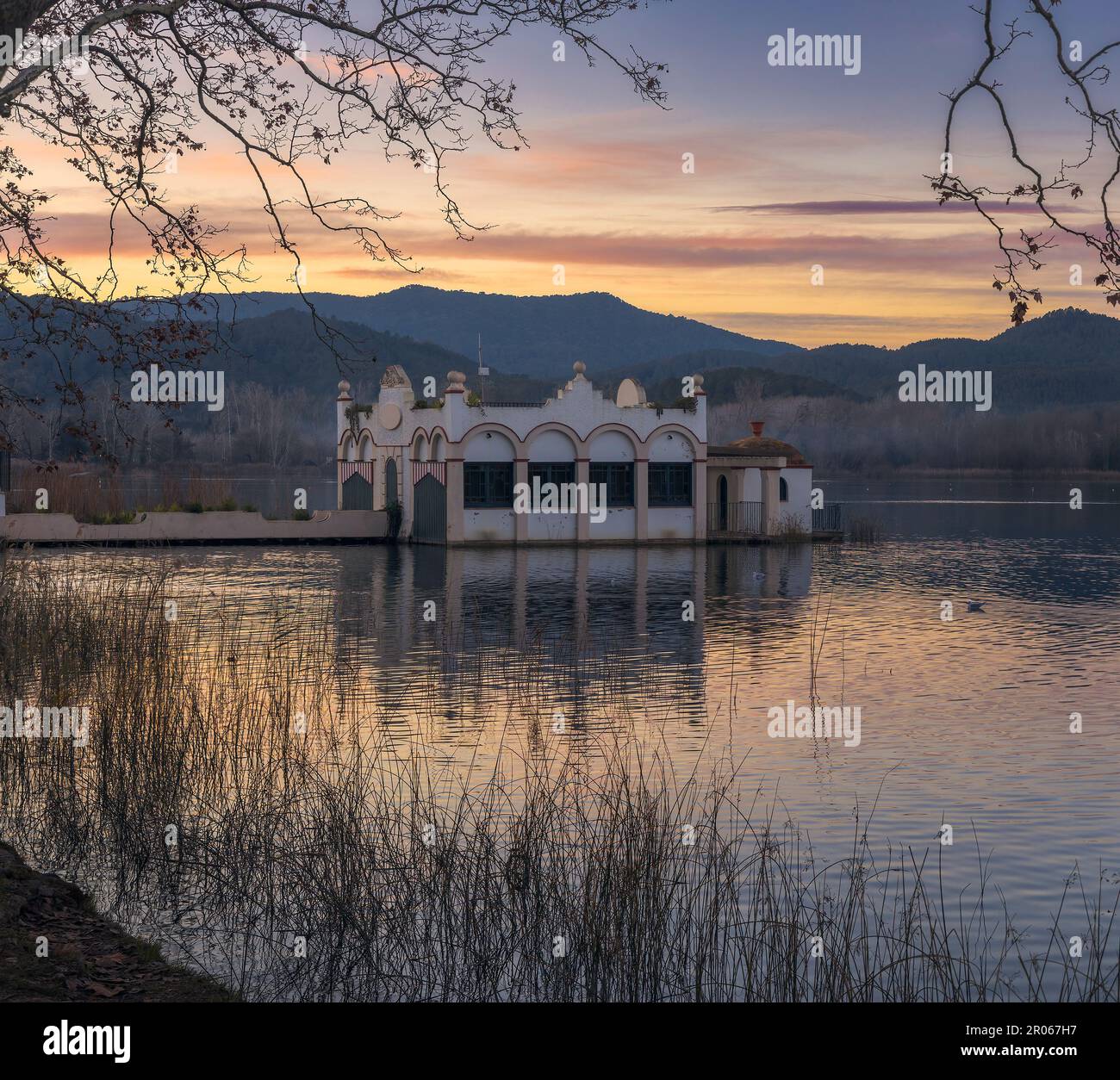La tranquilla bellezza del lago Banyoles si riflette nel cielo al tramonto, incorniciato da una maestosa struttura costruita e circondato da alberi. Foto Stock