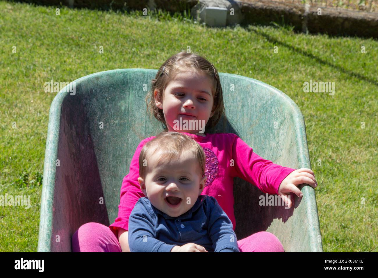 Immagine di due adorabili bambini sorridenti che si tengono all'interno di una carriola. Amore fraterno come giocano in giardino Foto Stock