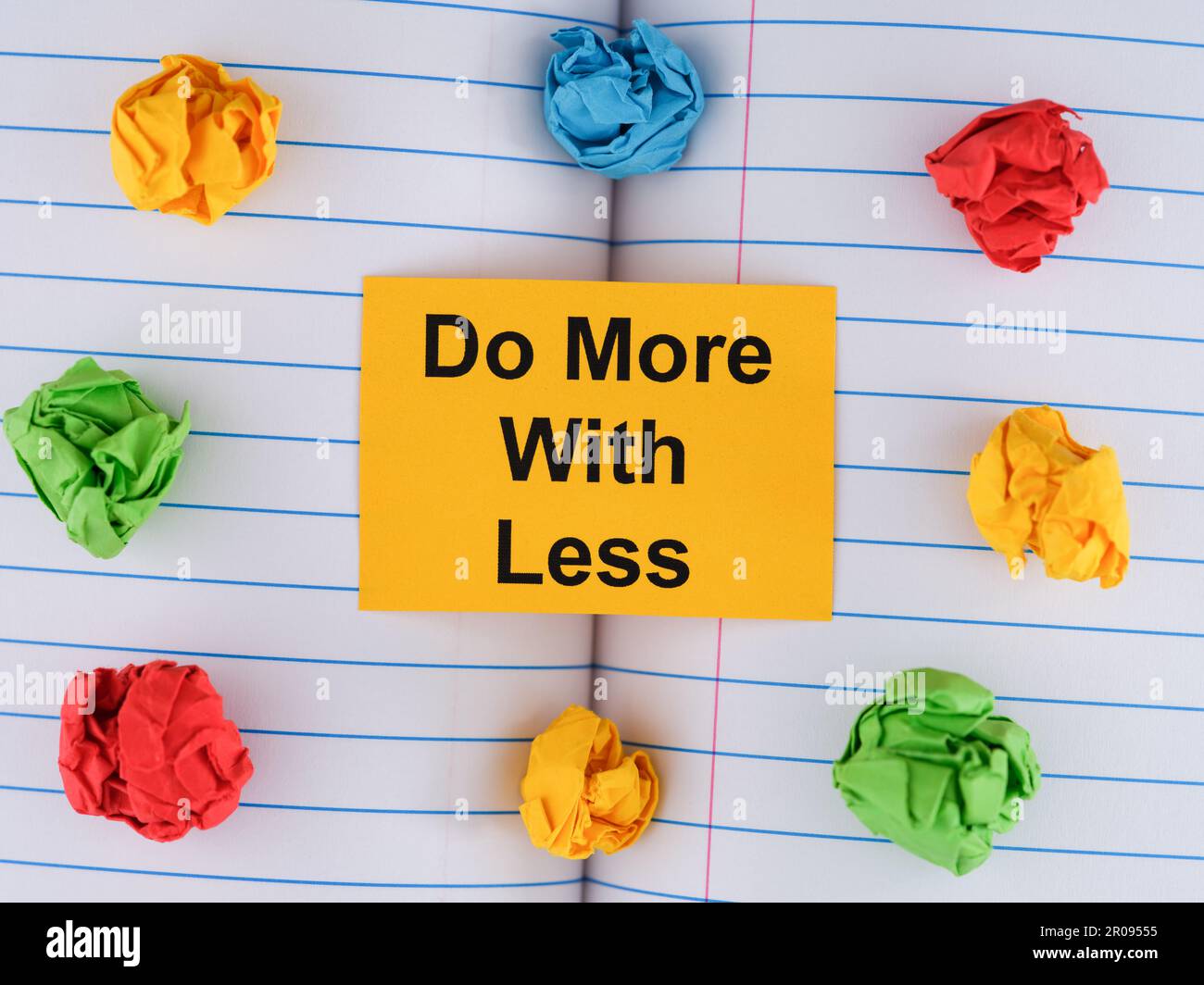 Una nota di carta gialla con la frase fare di più con meno su foglio di notebook con alcune palline di carta colorata stropicciata intorno ad esso. Primo piano. Foto Stock