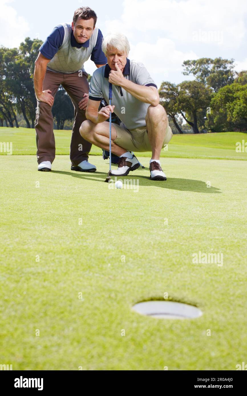 Ansiosa anticipazione del putt perfetto. Due uomini che guardano ansiosamente una palla da golf che rotola verso il buco sul verde. Foto Stock