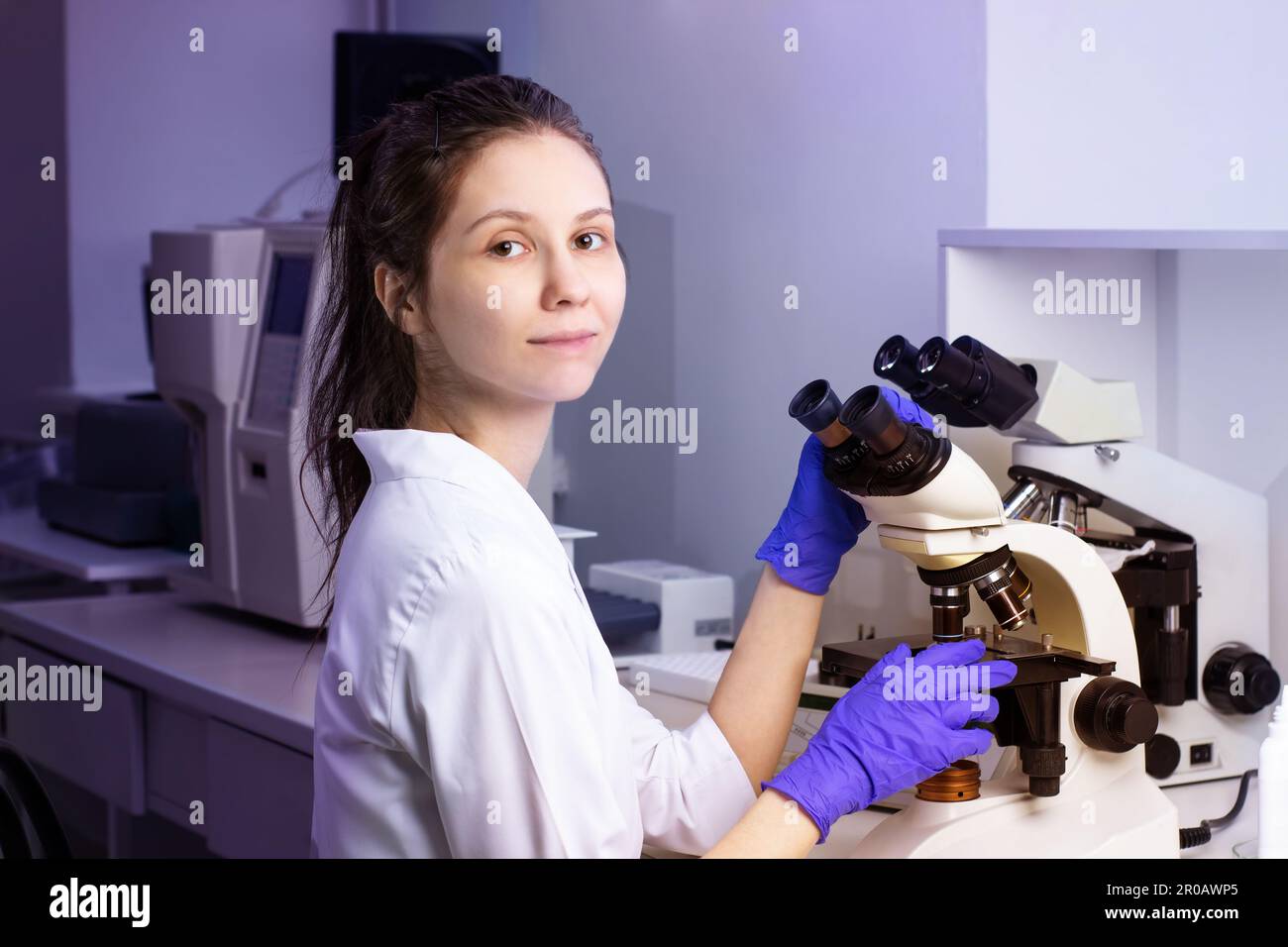 Una giovane donna medica (assistente di laboratorio) conduce uno studio di un campione di sangue su un microscopio. Circondato da attrezzature da laboratorio Foto Stock