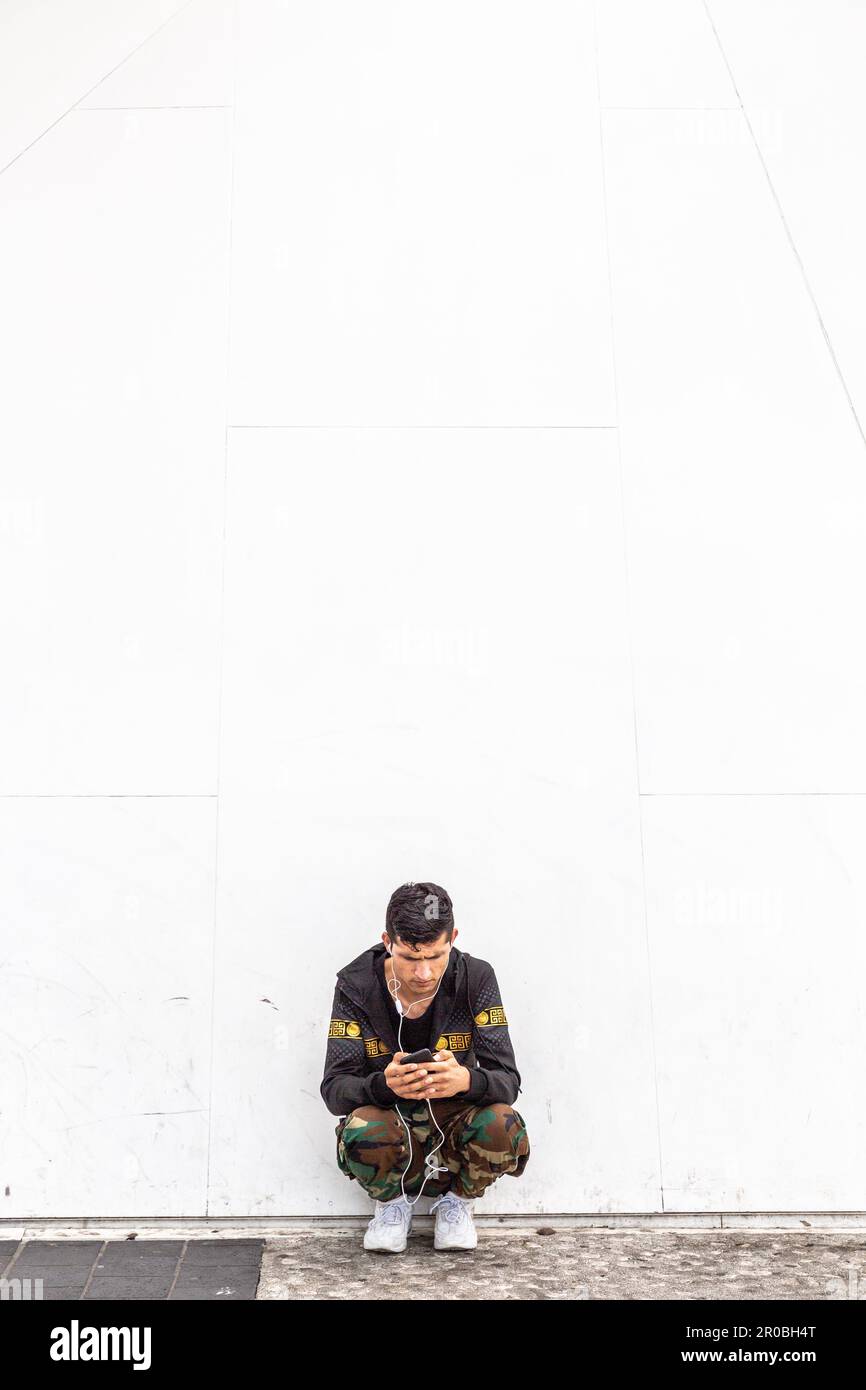 Giovane uomo che si rivolge al suo cellulare, accovacciandosi davanti a una parete bianca. Bruxelles. Foto Stock