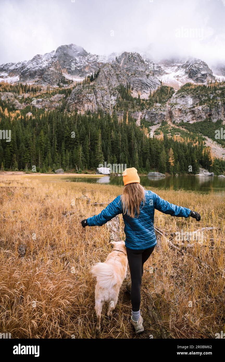 Ragazza escursionista e il suo cane accanto ad un lago alpino con larici Foto Stock