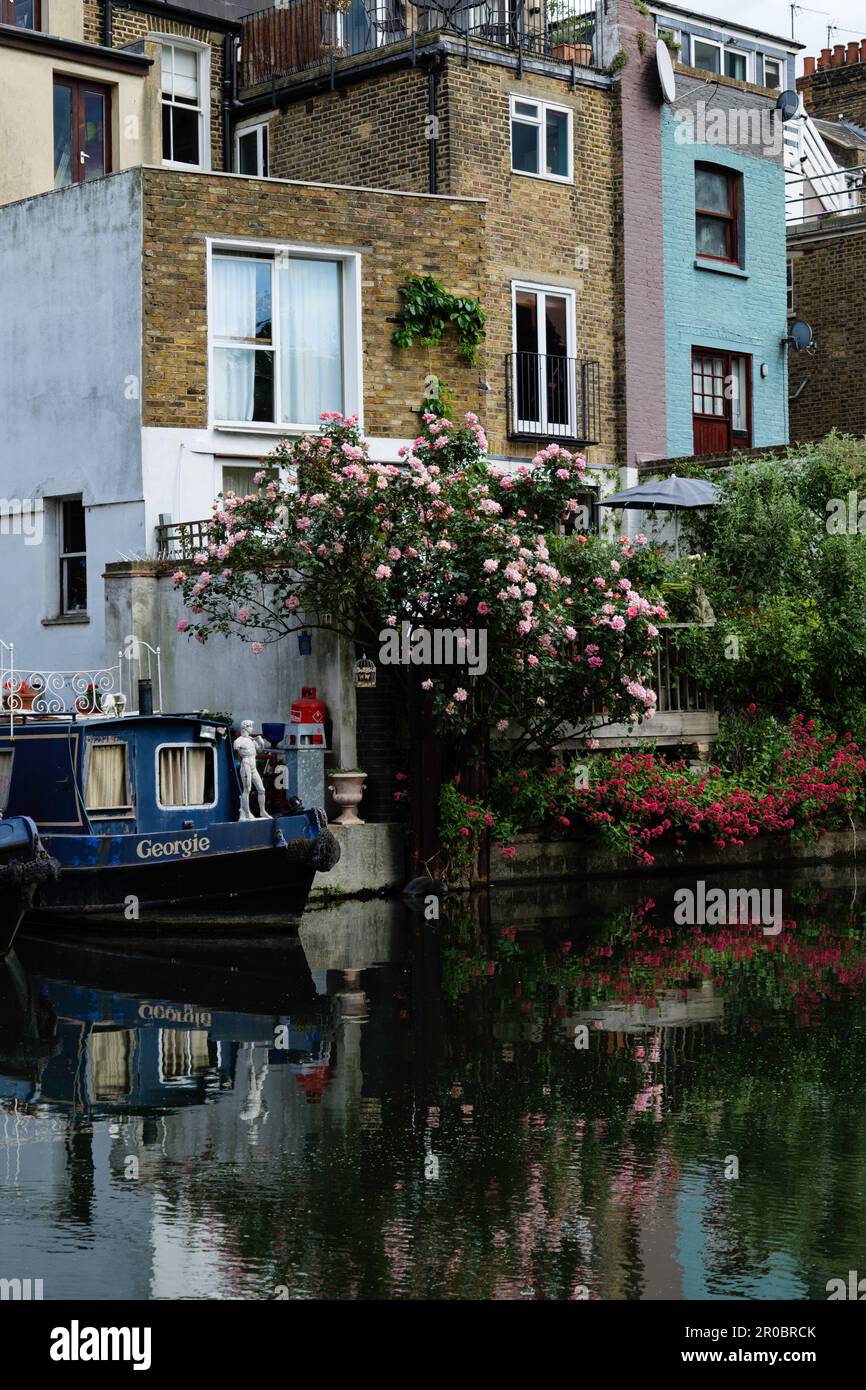 Londra - 05 21 2022: Casa galleggiante ormeggiato sul Canal Grande Union al largo di Elkstone Rd vicino a una casa e piante di rosa e geranio Foto Stock