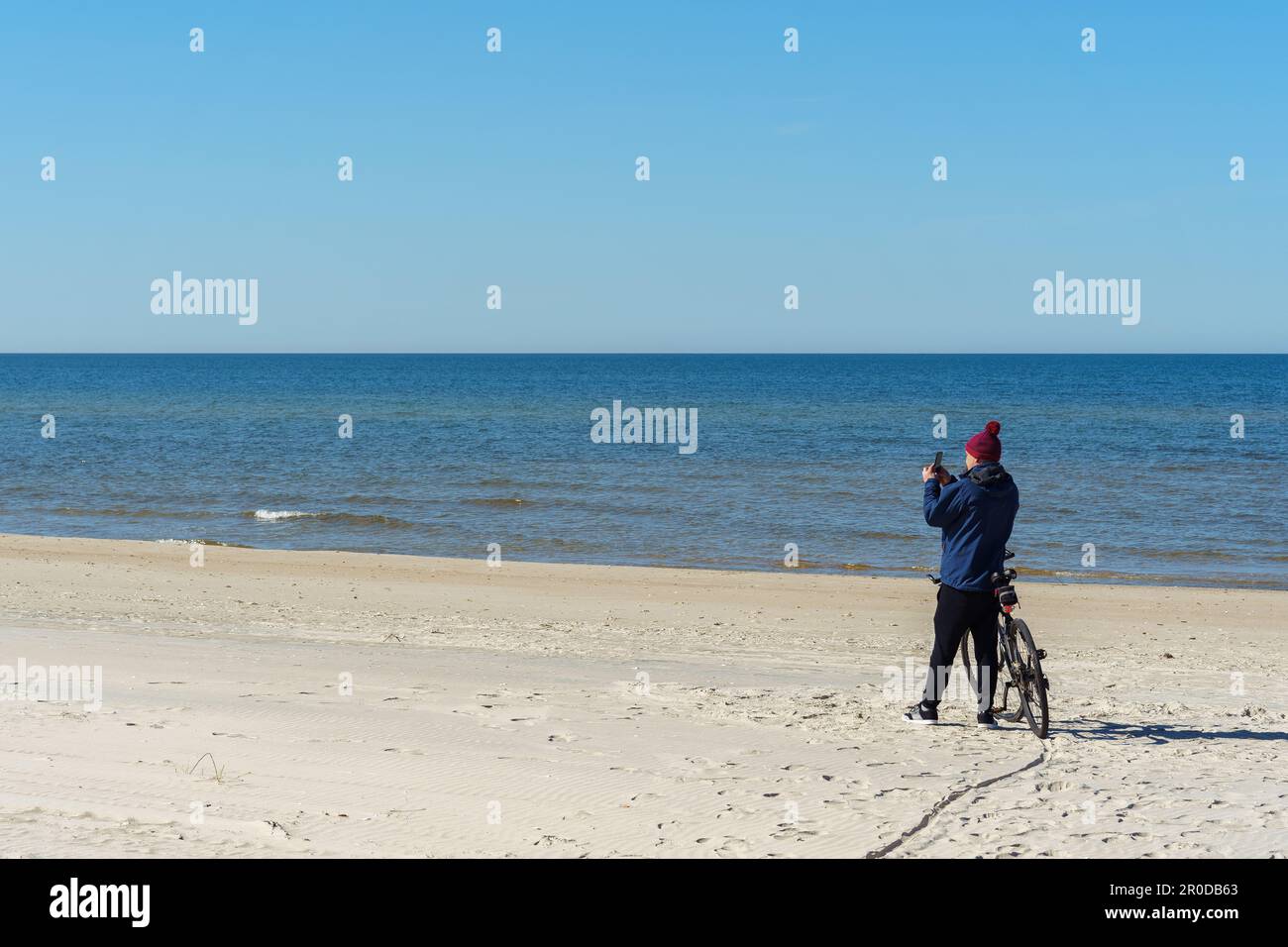 Un uomo in abiti caldi e un cappello scatta una foto sul suo telefono in piedi vicino alla sua bicicletta, viaggiando lungo il Mar Baltico sullo Spit Curoniano, Neringa, li Foto Stock