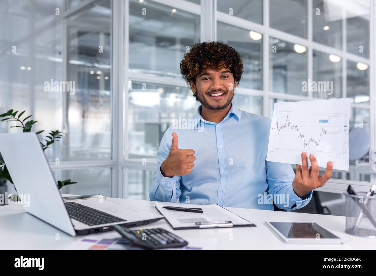 Ritratto di giovane finanziatore di successo documenti in ufficio, uomo sorridente e guardando la fotocamera mostra grafico con dinamiche positive dei profitti economici, uomo d'affari al lavoro con il computer portatile. Foto Stock