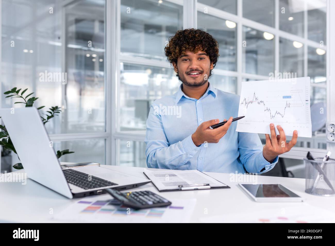 Ritratto di giovane finanziatore di successo documenti in ufficio, uomo sorridente e guardando la fotocamera mostra grafico con dinamiche positive dei profitti economici, uomo d'affari al lavoro con il computer portatile. Foto Stock