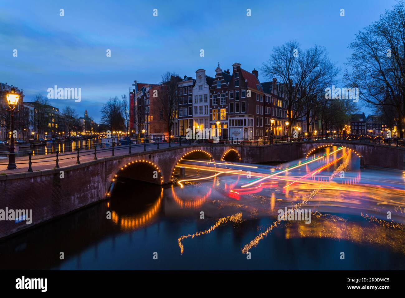 Ponti illuminati al crepuscolo ora blu ad Amsterdam, Olanda, Paesi Bassi nel mese di aprile lunga esposizione multipla con luci di barche che passano sotto il ponte Foto Stock