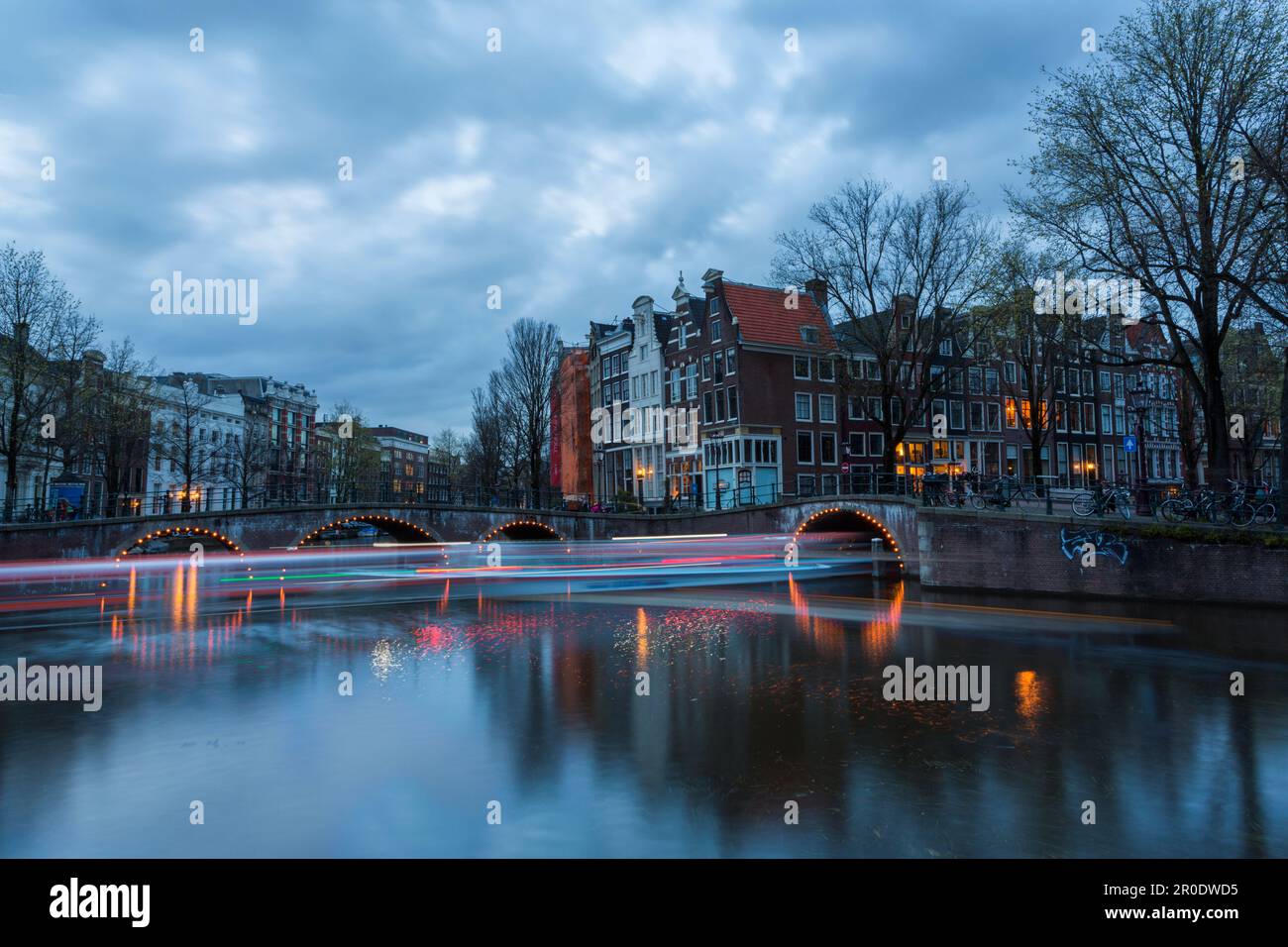 Ponti illuminati al crepuscolo ora blu ad Amsterdam, Olanda, Paesi Bassi nel mese di aprile lunga esposizione multipla con luci di barche che passano sotto il ponte Foto Stock