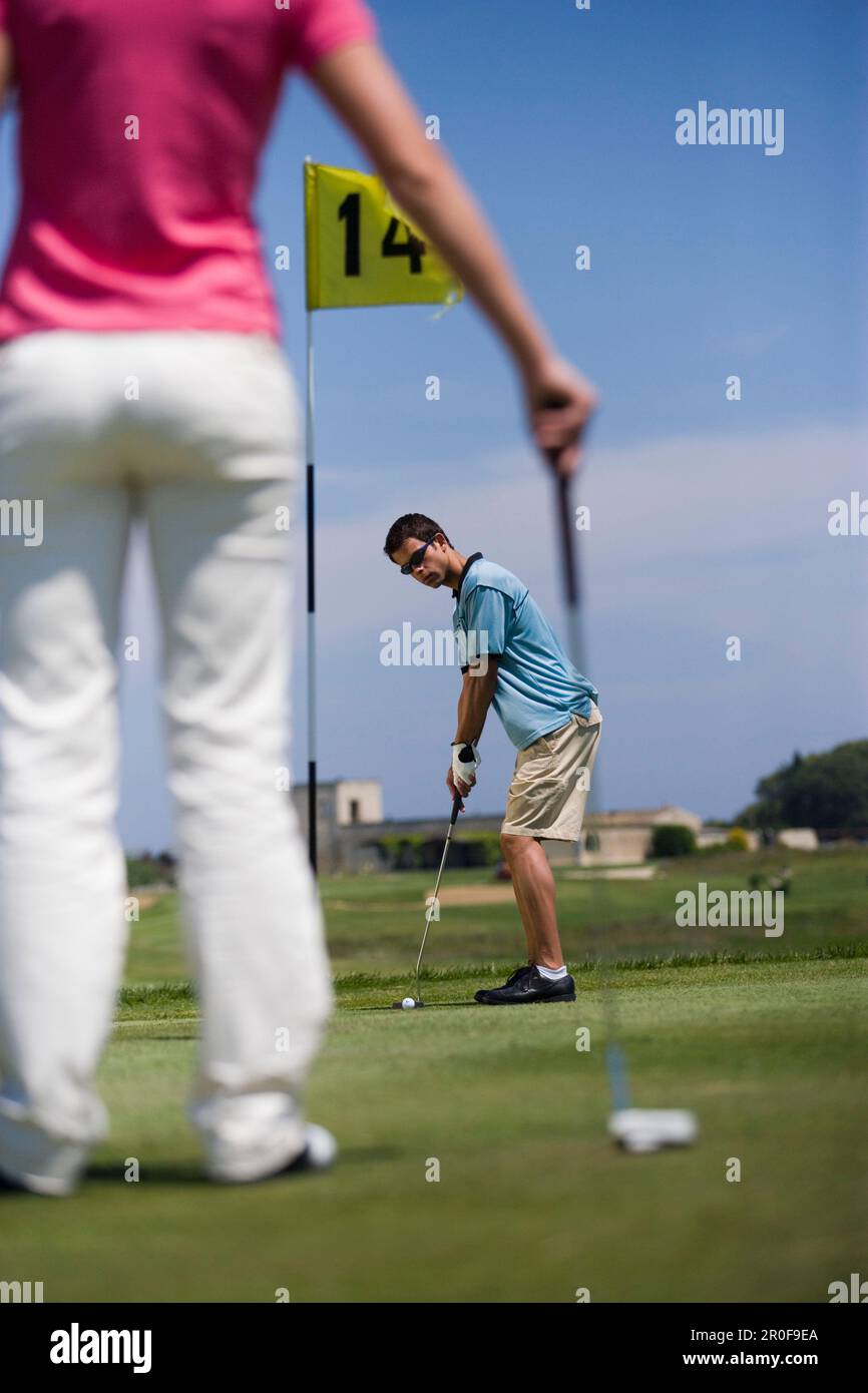 Golfista maschio che si prepara a colpire la palla, donna in piedi davanti, Puglia, Italia Foto Stock
