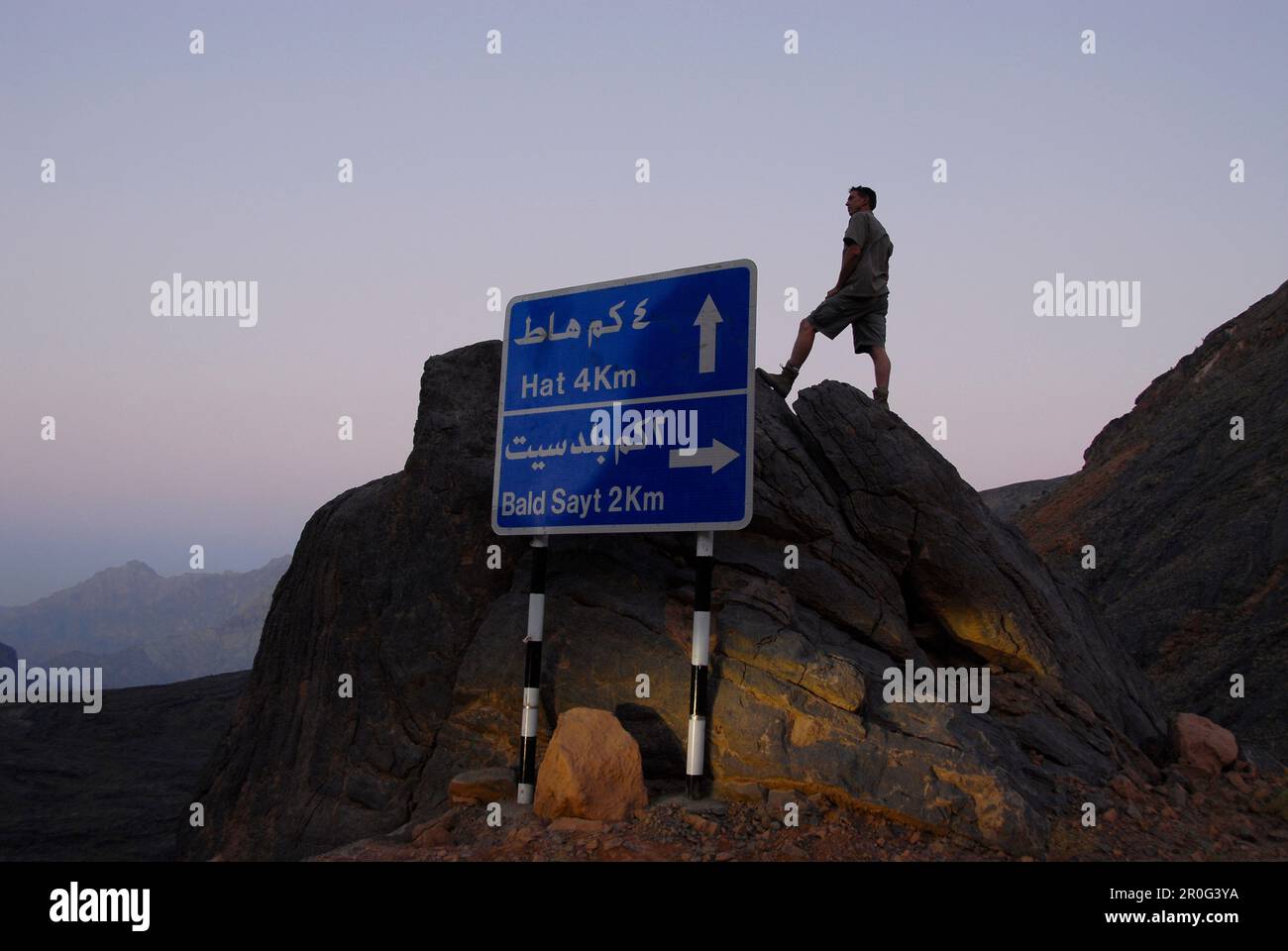 Un cartello stradale e un uomo su una roccia all'alba, al Hajar montagne, Oman, Asia Foto Stock