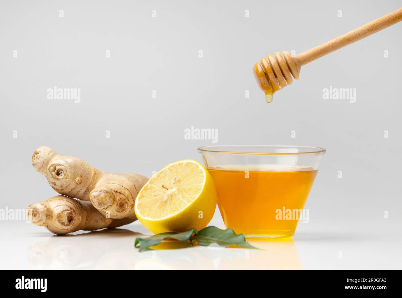 Miele in una ciotola di vetro, cucchiaia versando miele, zenzero e limone tagliato con foglie verdi su fondo bianco. Versando miele in un vaso, bastone di miele Foto Stock