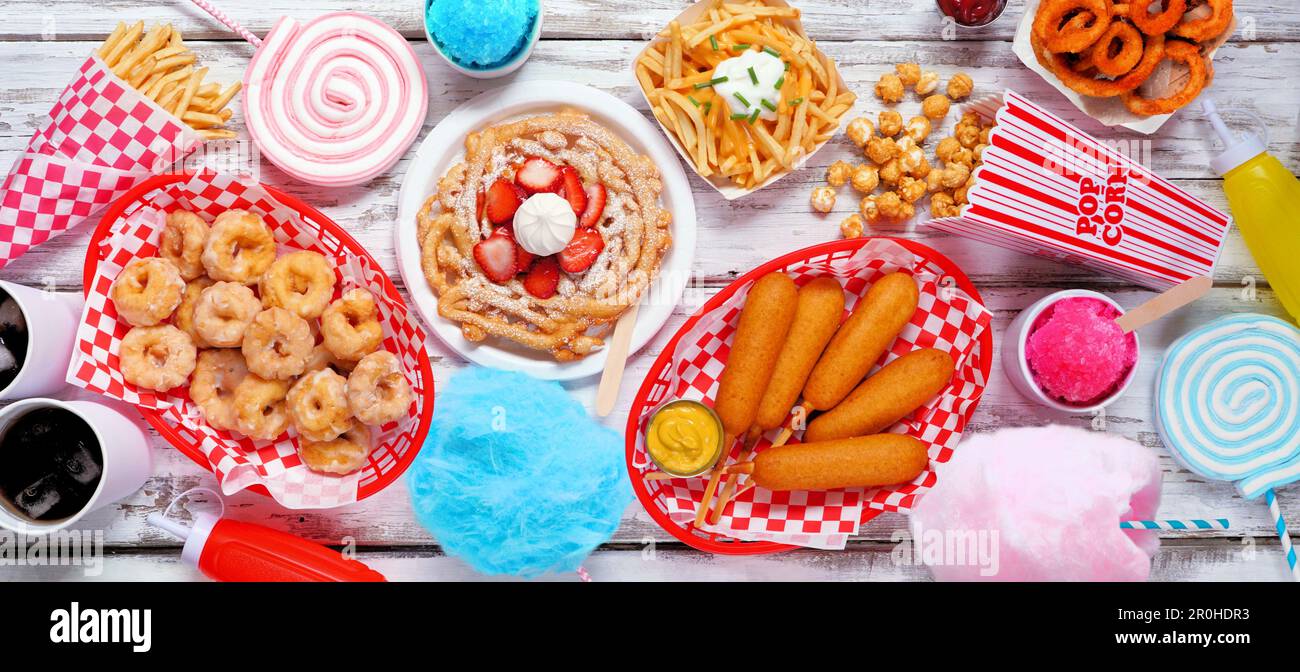 Scena del tavolo del cibo a tema di Carnevale su uno sfondo di bandiera di legno bianco. Vista dall'alto. Concetto di fiera estiva. Cani da granoturco, torta ad imbuto, caramelle di cotone e spuntini. Foto Stock