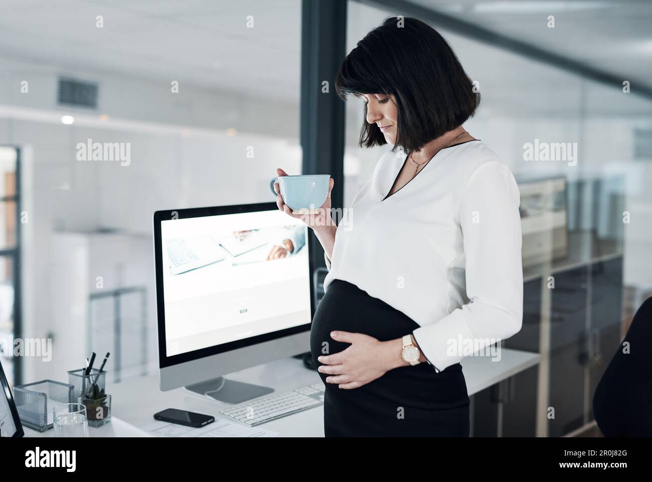 Im bere il vostro tè preferito. una donna d'affari incinta attraente che si alza da sola in ufficio e che ha una tazza di tè. Foto Stock