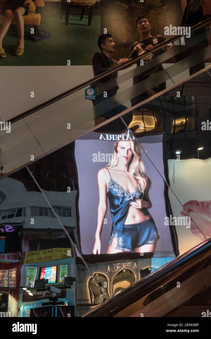 Mentre due giovani uomini cinesi in piedi su scala mobile un riflesso di una pubblicità illuminata con una bella signora bionda può essere visto, Kowloon, Hongkong, Cina, Asia Foto Stock