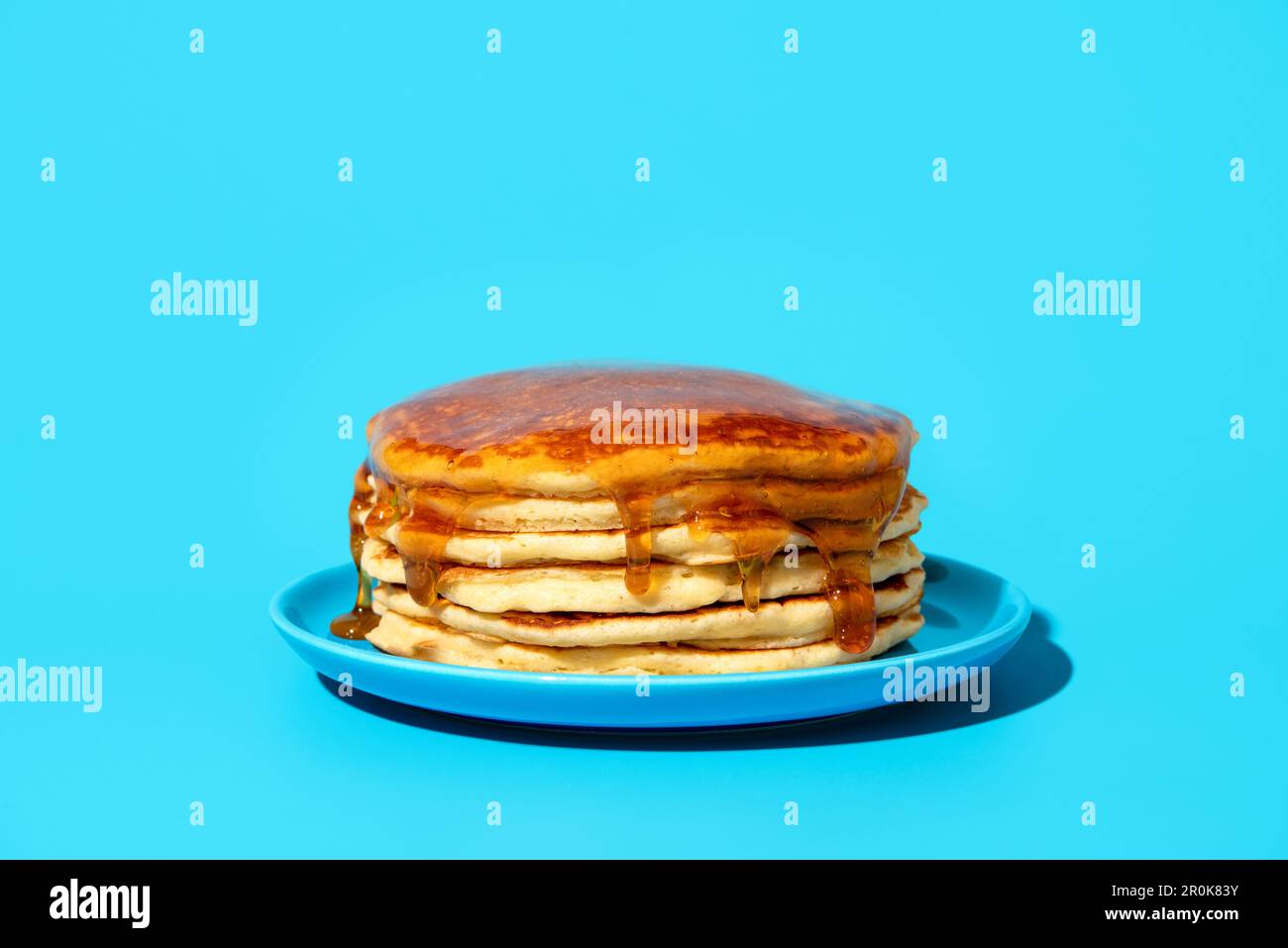 Pila di frittelle ricoperte di sciroppo su un piatto blu, minimalista su un tavolo blu. Deliziosi pancake fatti in casa con sciroppo d'acero in luce brillante su un co Foto Stock