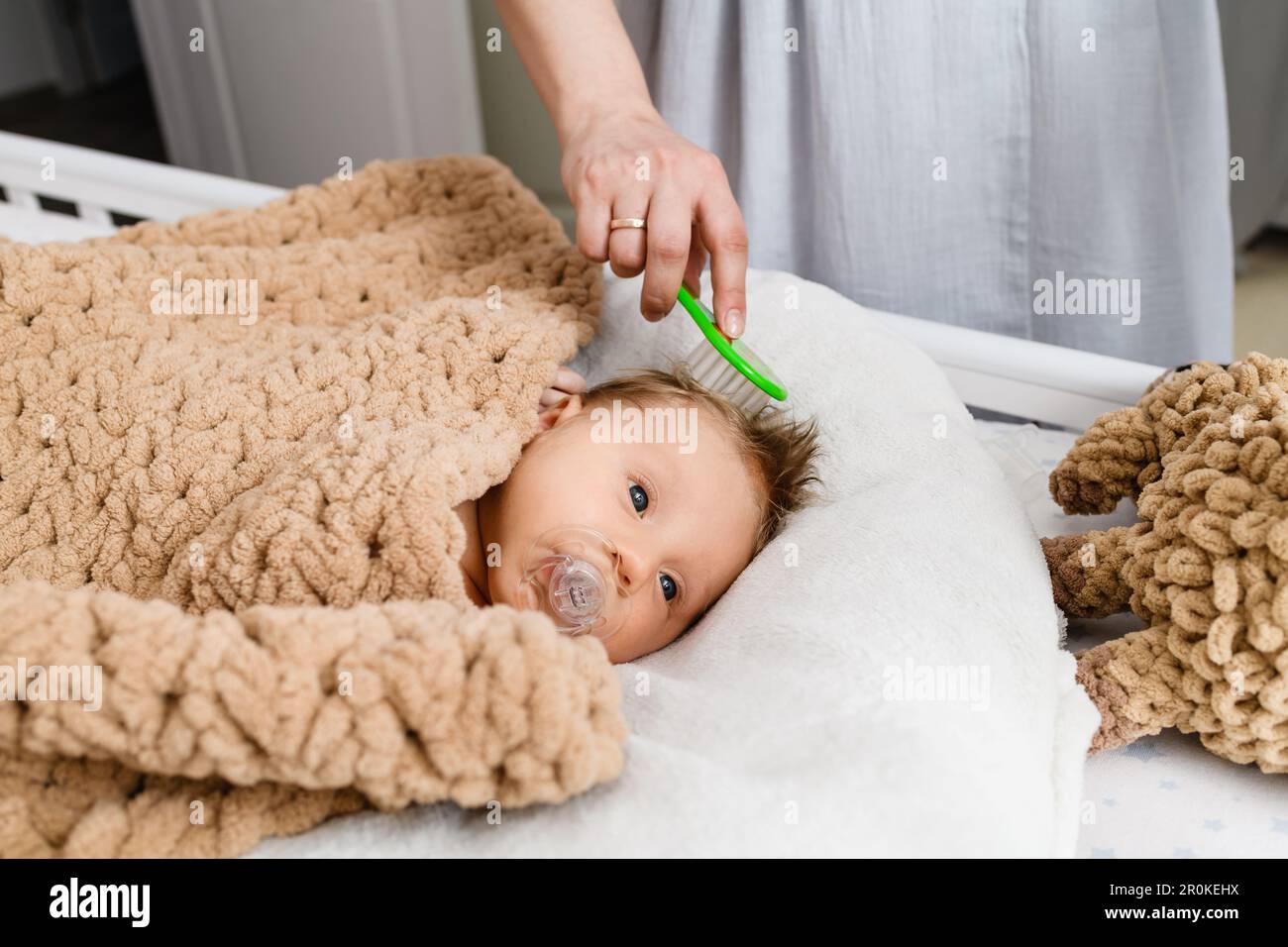 Spazzola a mano della madre i primi capelli del neonato, il bambino è adagiato in una culla coperta morbida e intima e succhiata, succhietto, piumino Foto Stock