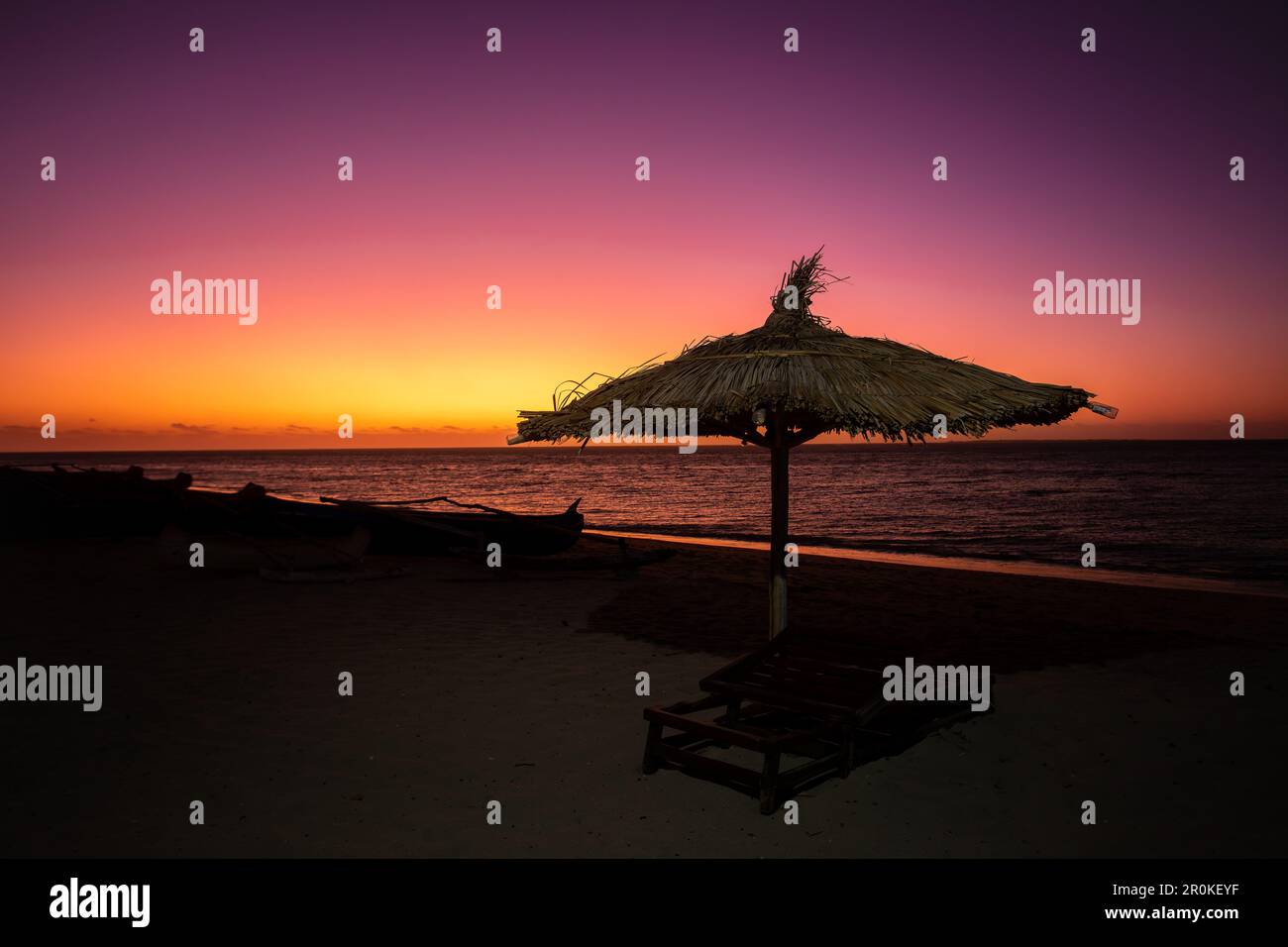 Cielo rosso vibrante al tramonto sulla spiaggia di Anakao in Madagascar, con un ombrellone tropicale e lettini, fuga perfetta dalla vita quotidiana. Foto Stock