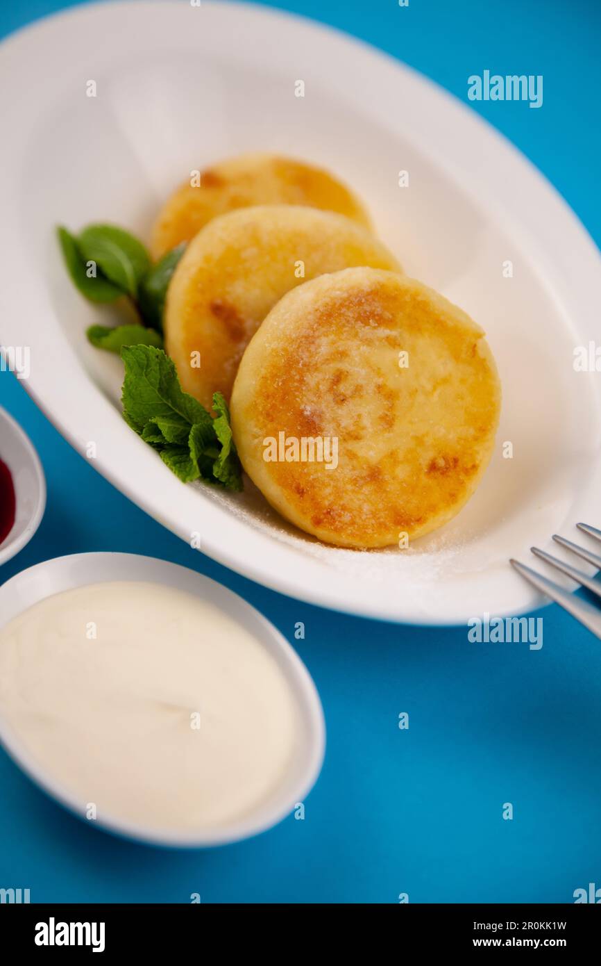 Frittelle di formaggio ricotta rotonda in un piatto bianco su sfondo blu. Cibo prima colazione. Cibo sano. Salsa di panna acida e menta. Primo piano vista dall'alto. Pasto dessert. Frittelle di ricotta al formaggio ricotta. Foto Stock