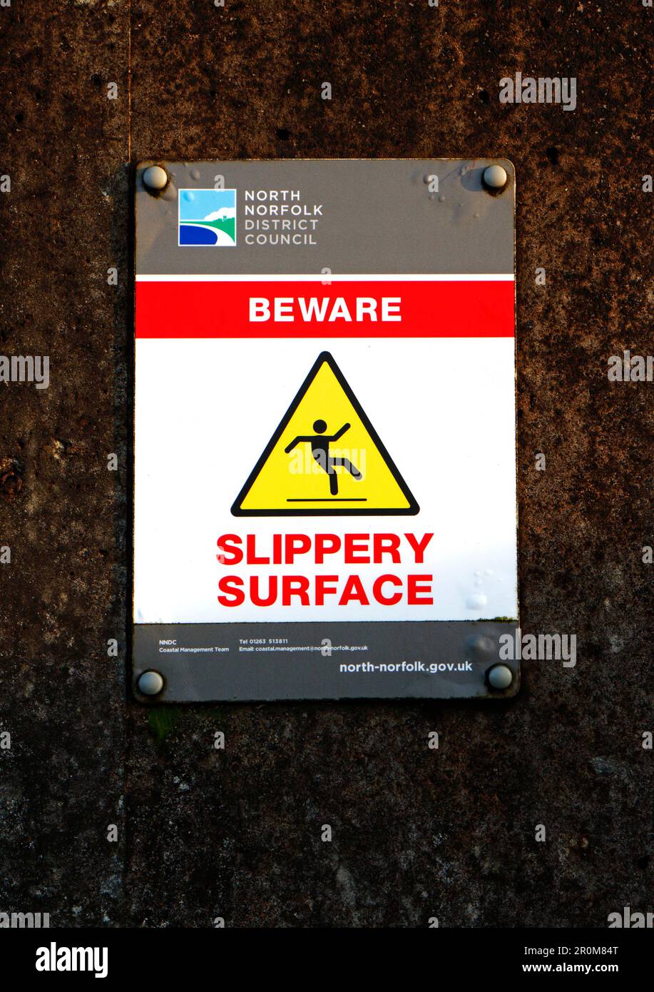 Un segno per fare attenzione alla superficie scivolosa sulle difese marine delle armature rocciose e all'area associata sulla costa nord del Norfolk a Sheringham, Norfolk, Inghilterra, Regno Unito. Foto Stock