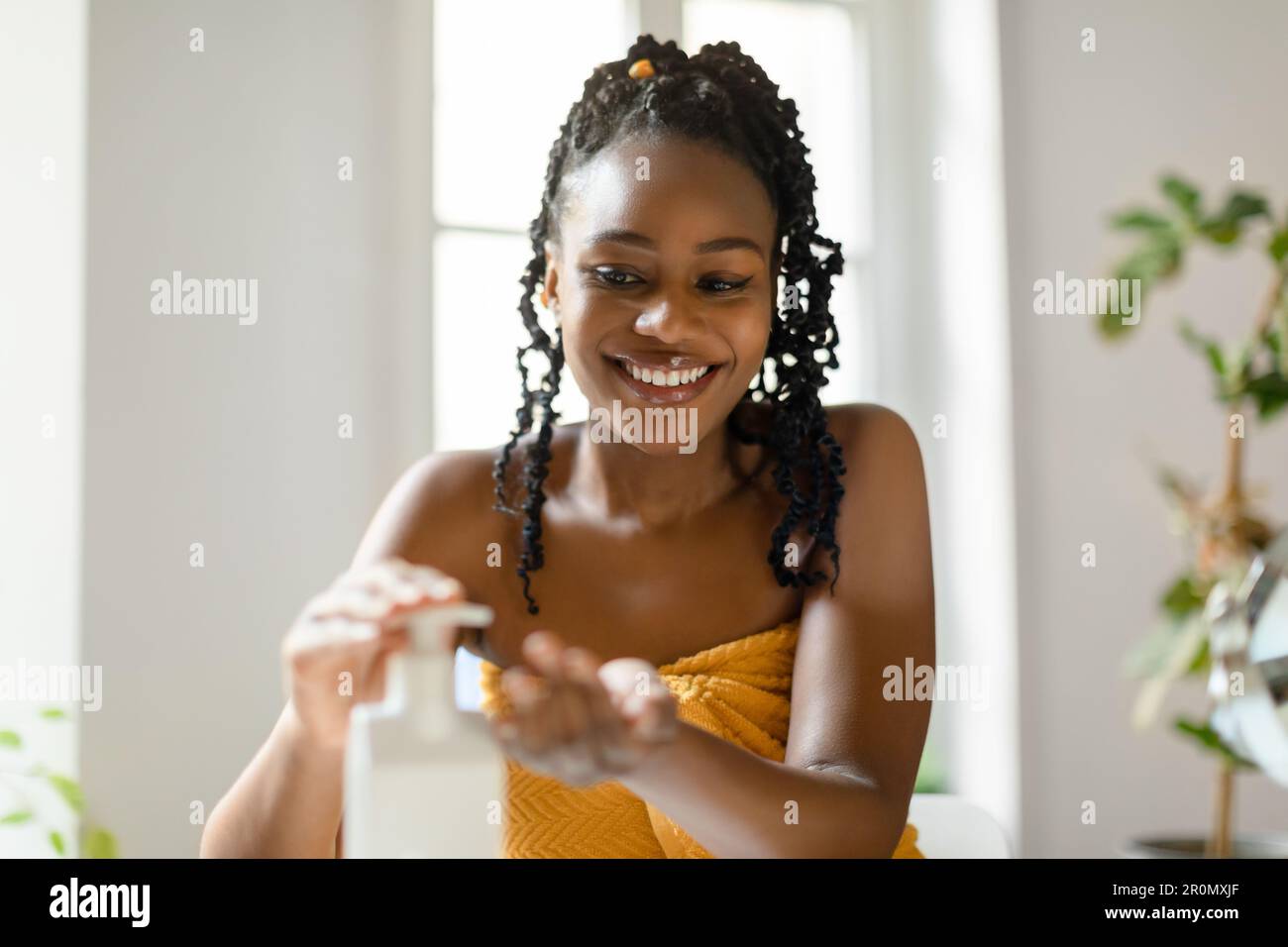 Giornata termale a casa. Donna nera felice versando lozione idratante del corpo o sapone liquido dalla bottiglia a portata di mano in bagno Foto Stock