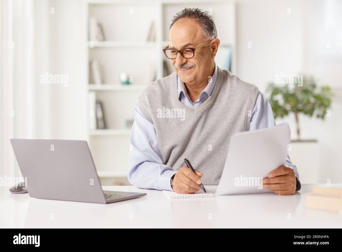 Uomo maturo che guarda un computer portatile a casa e scrive su un foglio Foto Stock