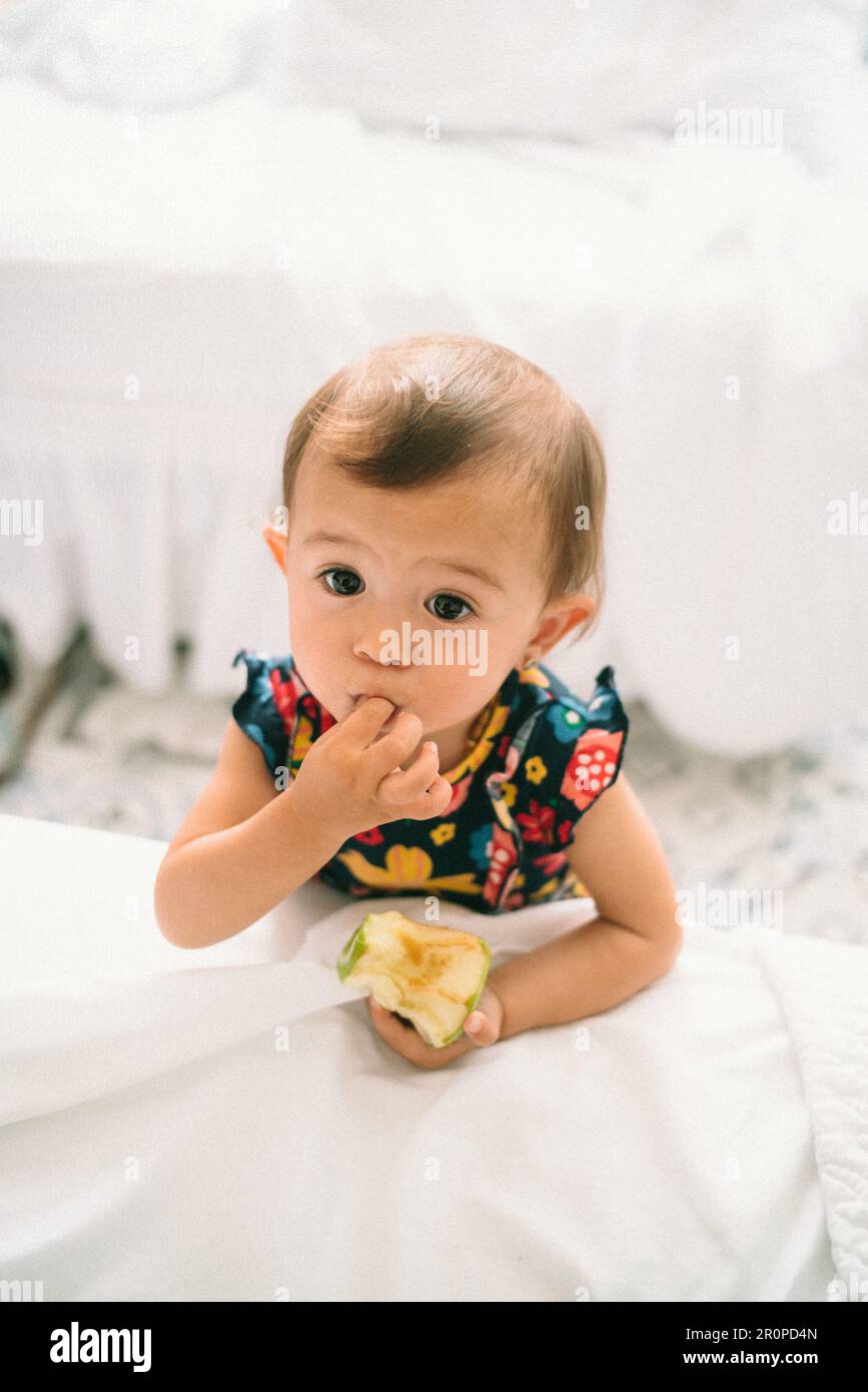 Una bambina cinese sorridente che indossa un abito floreale sta mangiando una mela con il dito in bocca Foto Stock