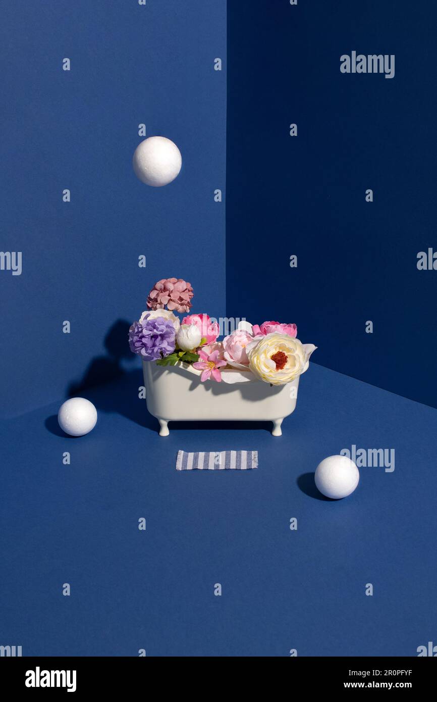 Vasca da bagno piena di fiori con bolle bianche in angolo di pareti blu. Concetto creativo minimo. Foto Stock