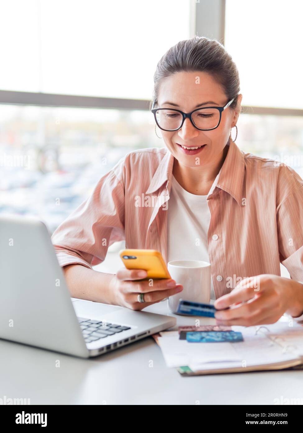 La donna sorridente lavora con un computer portatile e uno smartphone. La donna sta controllando le carte di credito o la carta d'identità. Ufficio moderno con finestre panoramiche. Centro di co-working Foto Stock