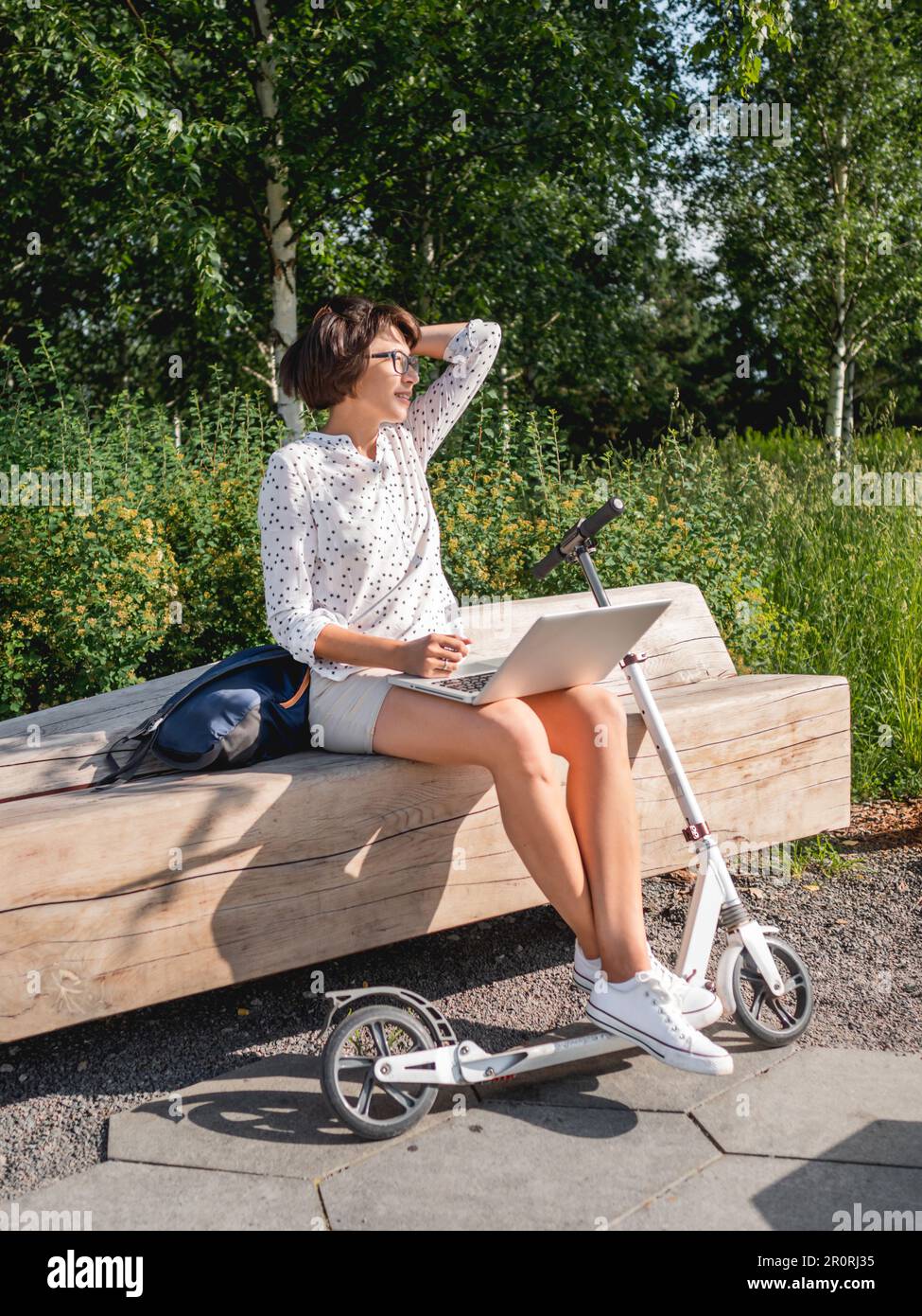 Donna seduta con un notebook sulla panca del parco urbano. Libero professionista al lavoro. Lo studente impara in remoto dall'esterno. Stile di vita moderno. Vibrazioni estive. All'aperto. Foto Stock