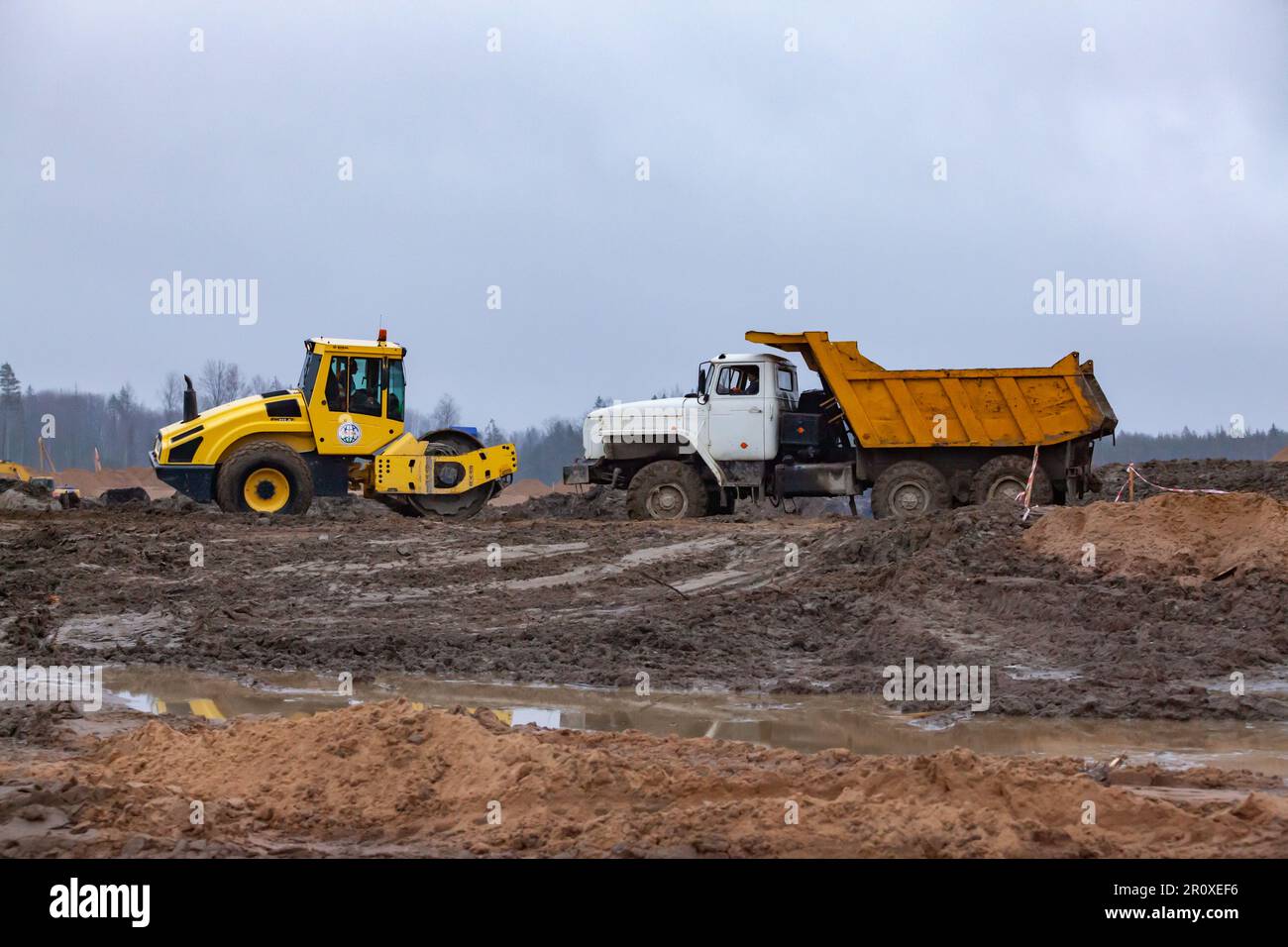 Ust-Luga, Leningrado oblast, Russia - 16 novembre 2021: Compattatore per asfalto e camion di scarico nel fango. Cantiere, lavori di fondo Foto Stock