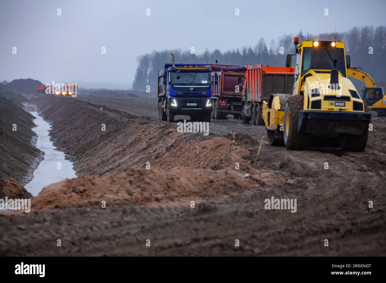 Ust-Luga, Leningrado oblast, Russia - 16 novembre 2021: Traffico su strada fangosa di cantiere in caso di pioggia. Trincea con acqua a sinistra Foto Stock