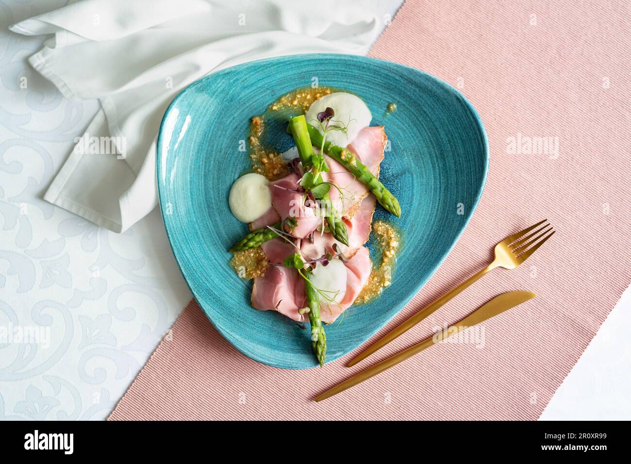 Vista dall'alto di un antipasto con asparagi, taleggio, arachidi e prosciutto al rosmarino su piatto blu. Tavolo da ristorante con stoffa rosa, tovagliolo bianco, posate dorate. Foto Stock