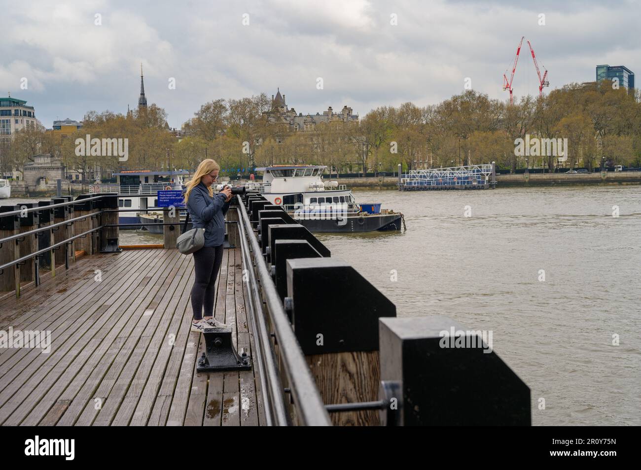 Una donna bionda fotografa cattura il vivace paesaggio urbano di Londra da un molo di legno lungo il Tamigi. Foto Stock