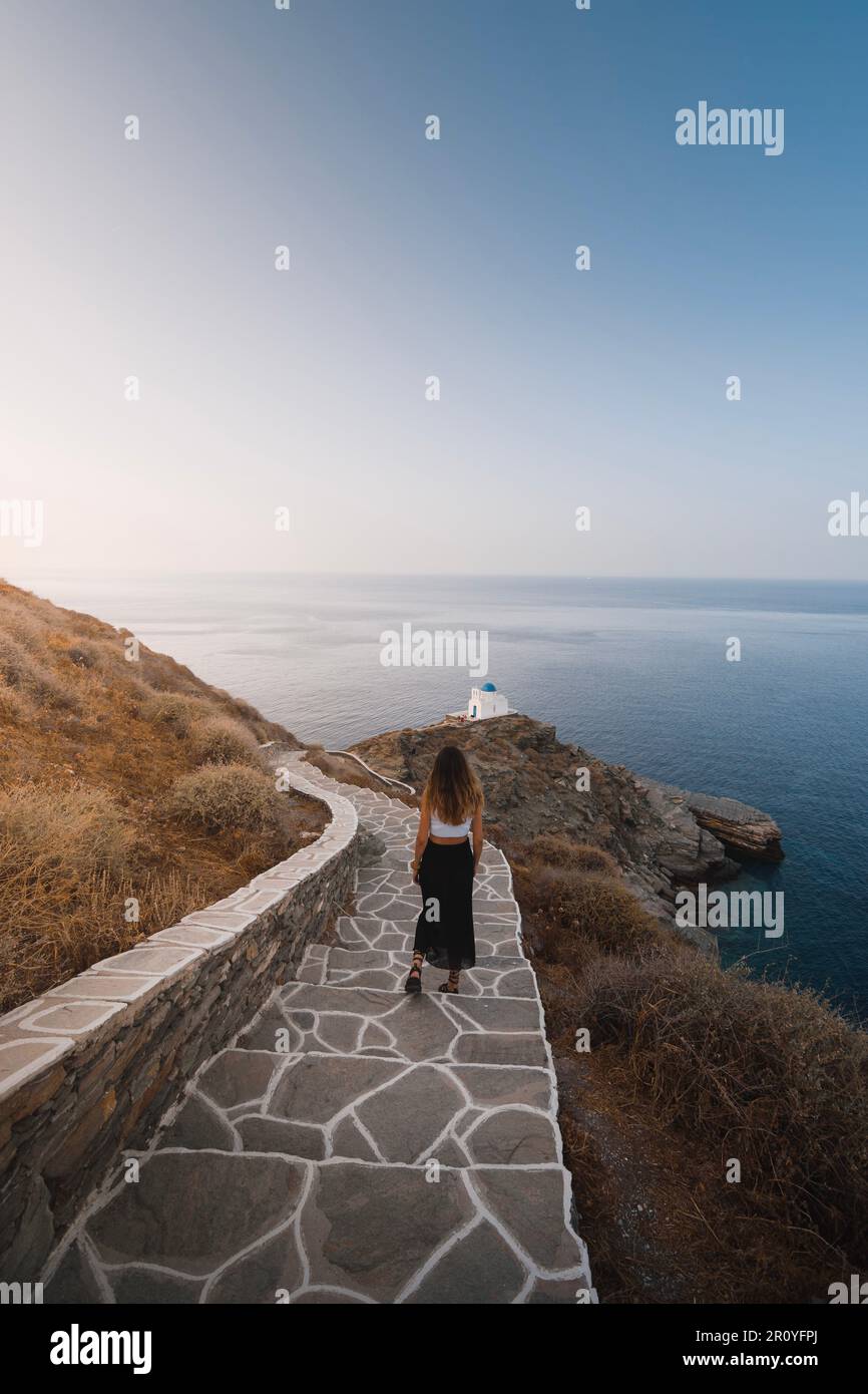 Giovane donna che cammina giù alcune graziose scale sulla strada per una chiesa sulla scogliera sull'isola greca di Sifnos durante il suo viaggio alle isole Cicladi in Foto Stock