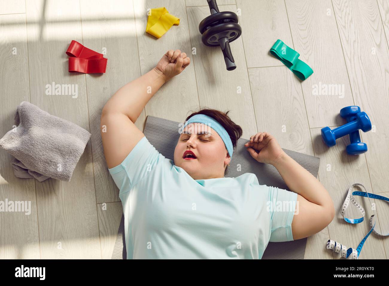 Divertente stanco pigro chubby donna che dorme sul pavimento tra le attrezzature per l'allenamento sportivo Foto Stock
