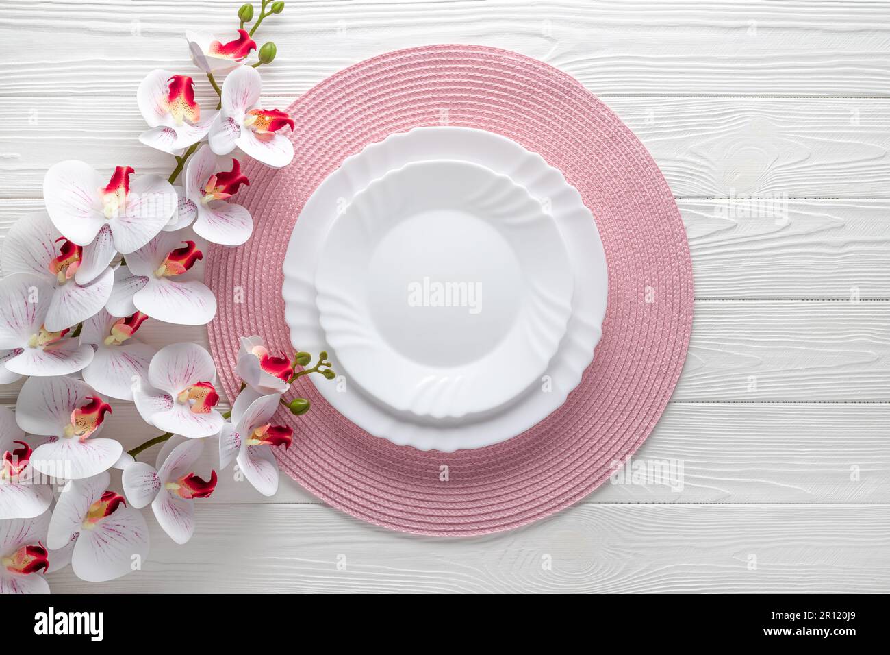 Ambiente festivo con tovagliolo rosa e fiori di orchidea. Piastre vuote su tavolo di legno bianco. Vista dall'alto. Cena festiva. Scheda o modello di menu, piatto d Foto Stock