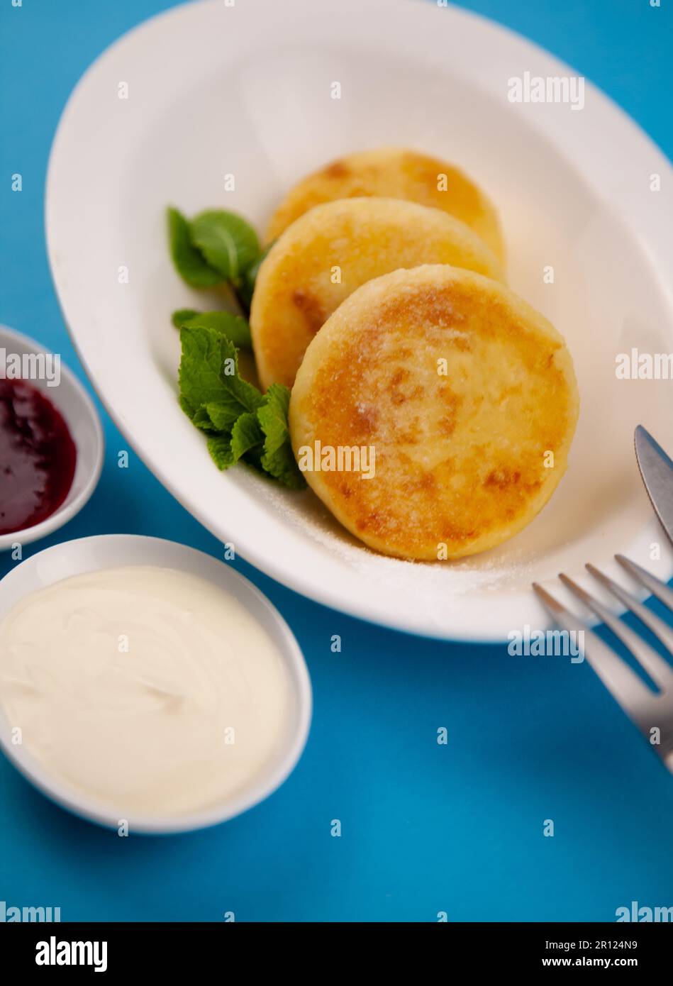 Frittelle di formaggio ricotta rotonda in un piatto bianco su sfondo blu. Cibo prima colazione. Cibo sano. Salsa di panna acida e menta. Primo piano vista dall'alto. Pasto dessert. Frittelle di ricotta al formaggio ricotta. Foto Stock