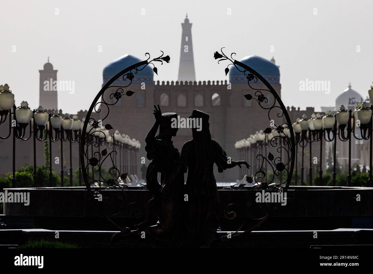 una silhouette statua di due figure con una ghirlanda incornicia la vista del minareto islam khoja e le porte della città murata di khiva uzbekistan Foto Stock