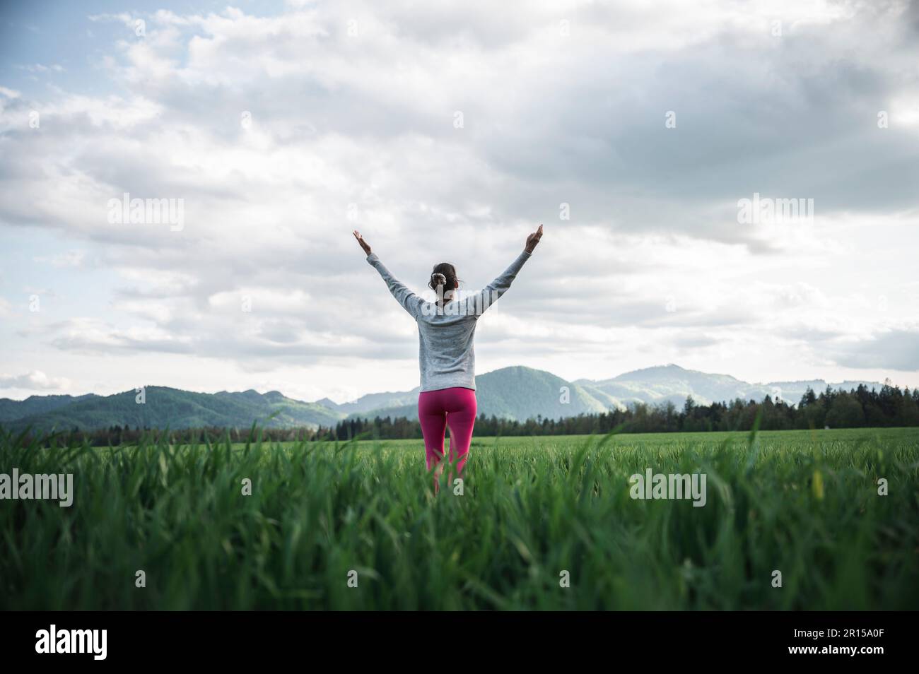 Vista posteriore ad angolo basso di una donna in piedi nel mezzo di un campo verde o prato godendo la vita e meditando con le braccia sollevate in alto. Sotto sp. Nuvoloso Foto Stock