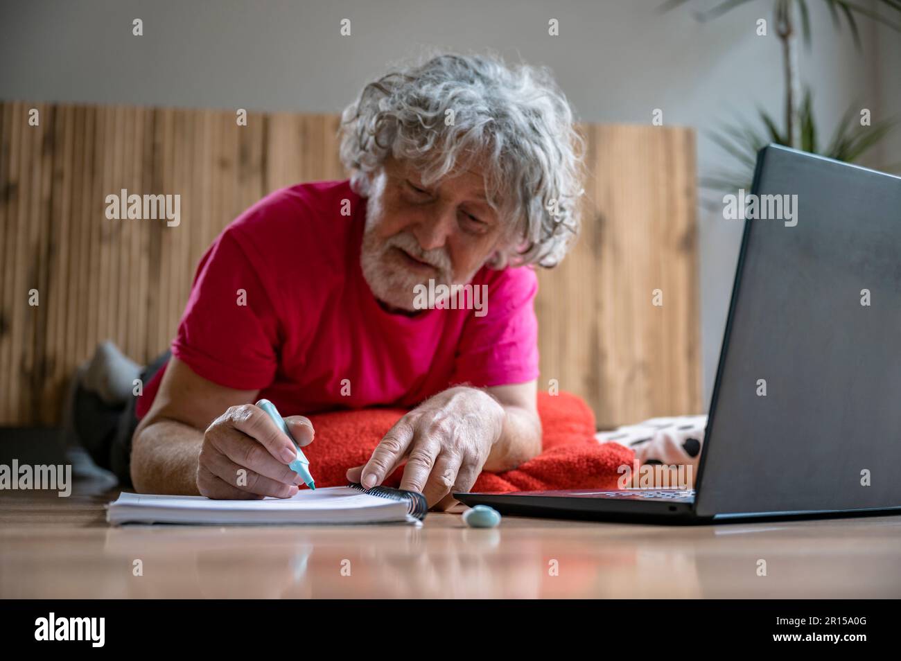 Uomo anziano che si trova sul pavimento del soggiorno prendendo appunti nel suo notebook mentre frequenta una lezione online utilizzando un computer portatile. Foto Stock