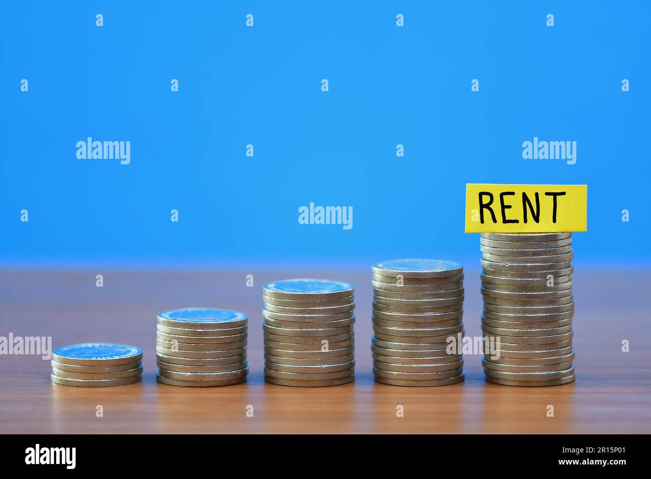 Una fila di una crescente pila di monete che illustra il crescente costo di affitto di famiglia a causa del crescente costo della vita, su uno sfondo blu Foto Stock