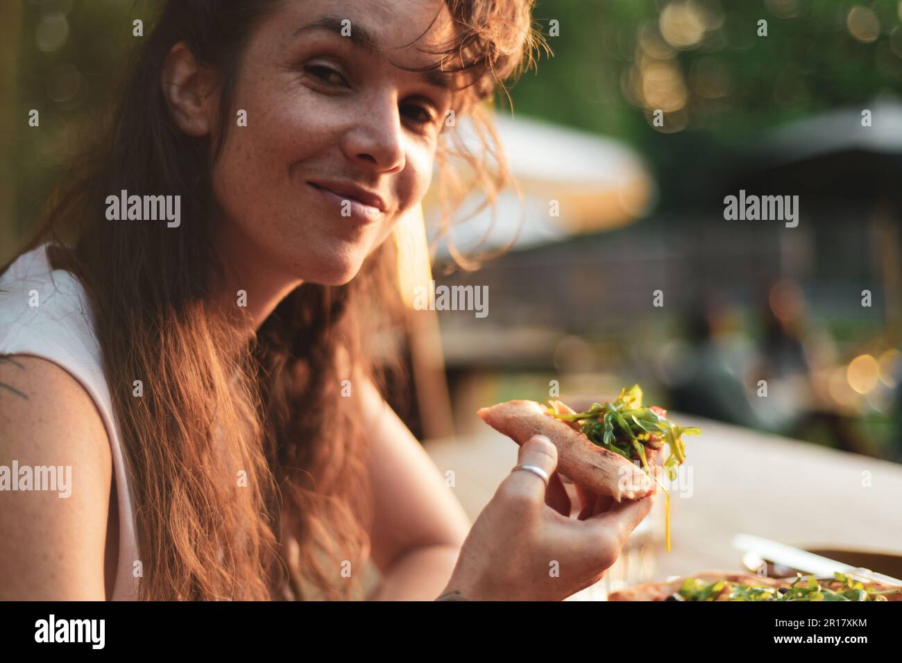 la donna sorride mentre mangia la pizza organica nel sole dorato fuori del caffè Foto Stock